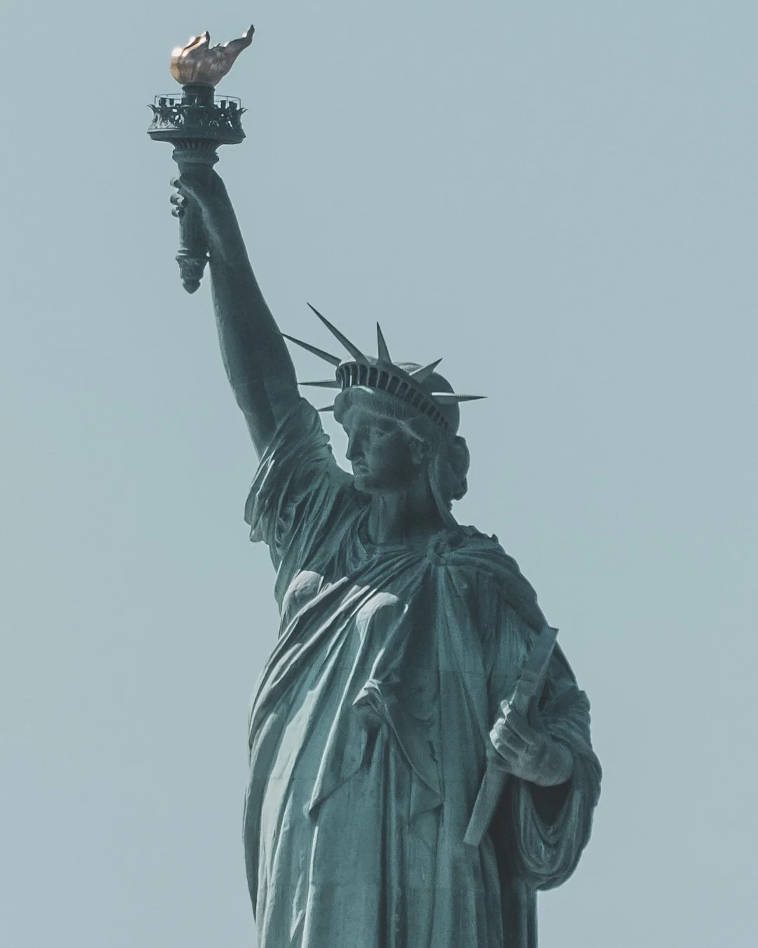 Close-up of the Statue of Liberty's face, crown, and torch, with a clear sky in the background. NEW YORK CITY
