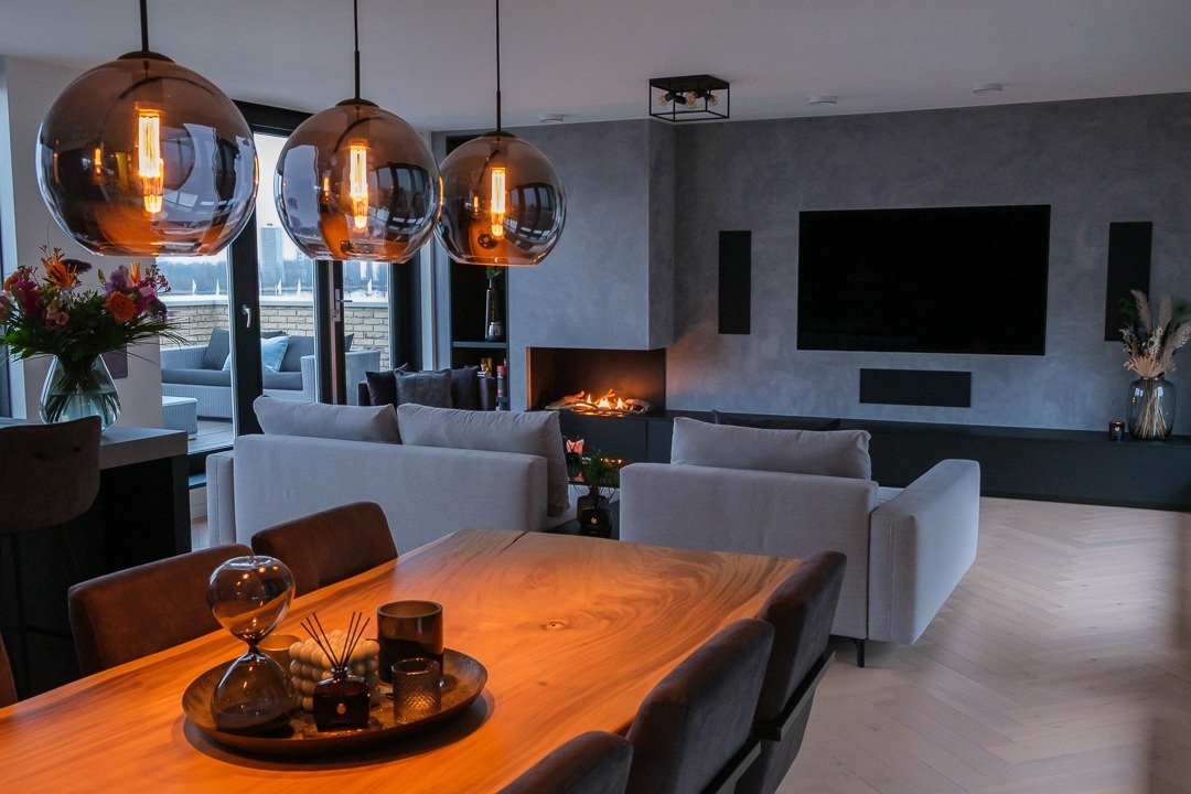 Modern living room with three hanging pendant lights, a TV on the wall, a mounted fireplace, a gray sectional sofa, and a wooden dining table with chairs. There are large windows and sliding doors leading to an outdoor seating area.
