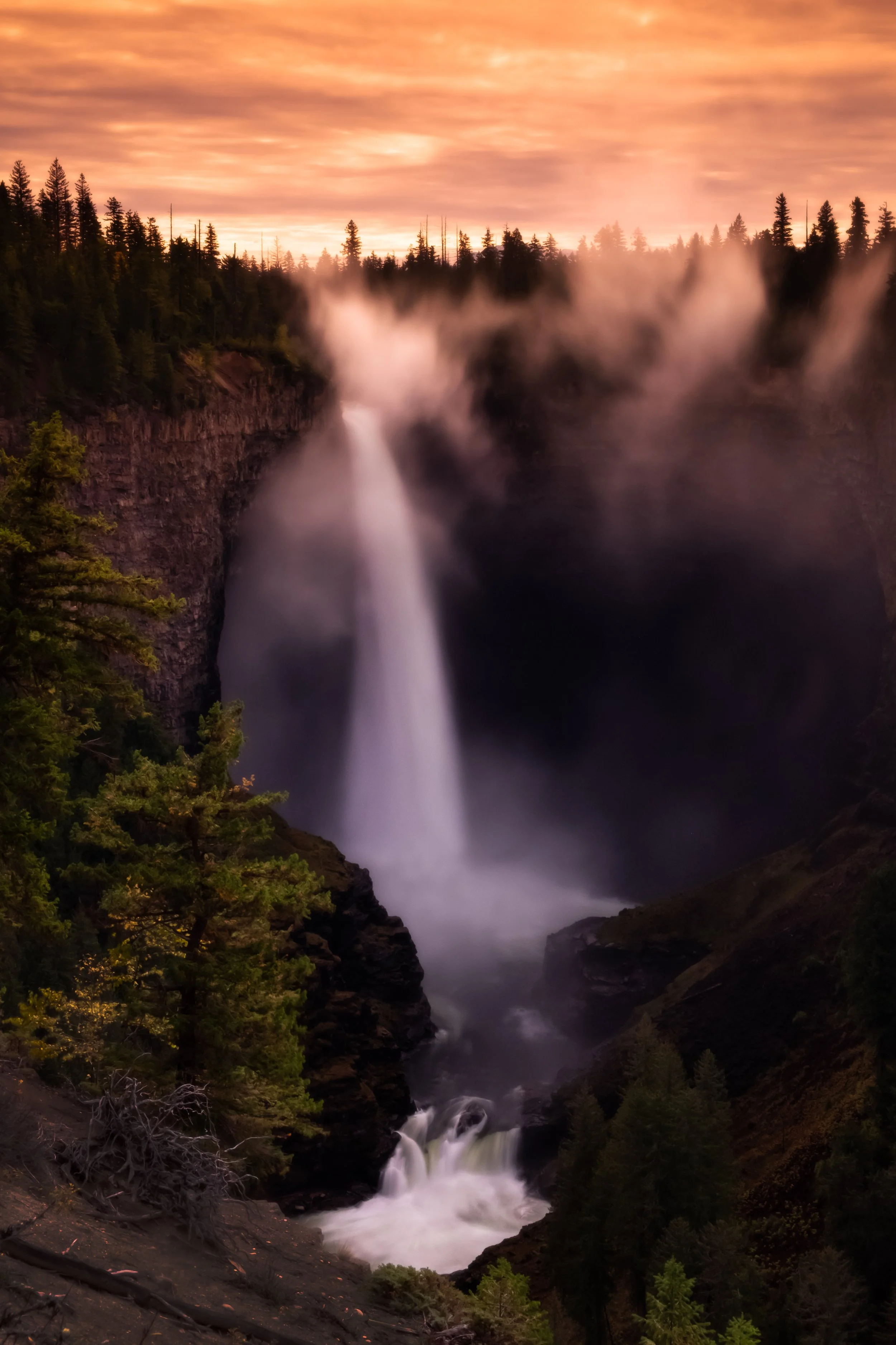 A waterfall cascading down a cliff surrounded by pine trees during sunset with colorful sky and mist.
