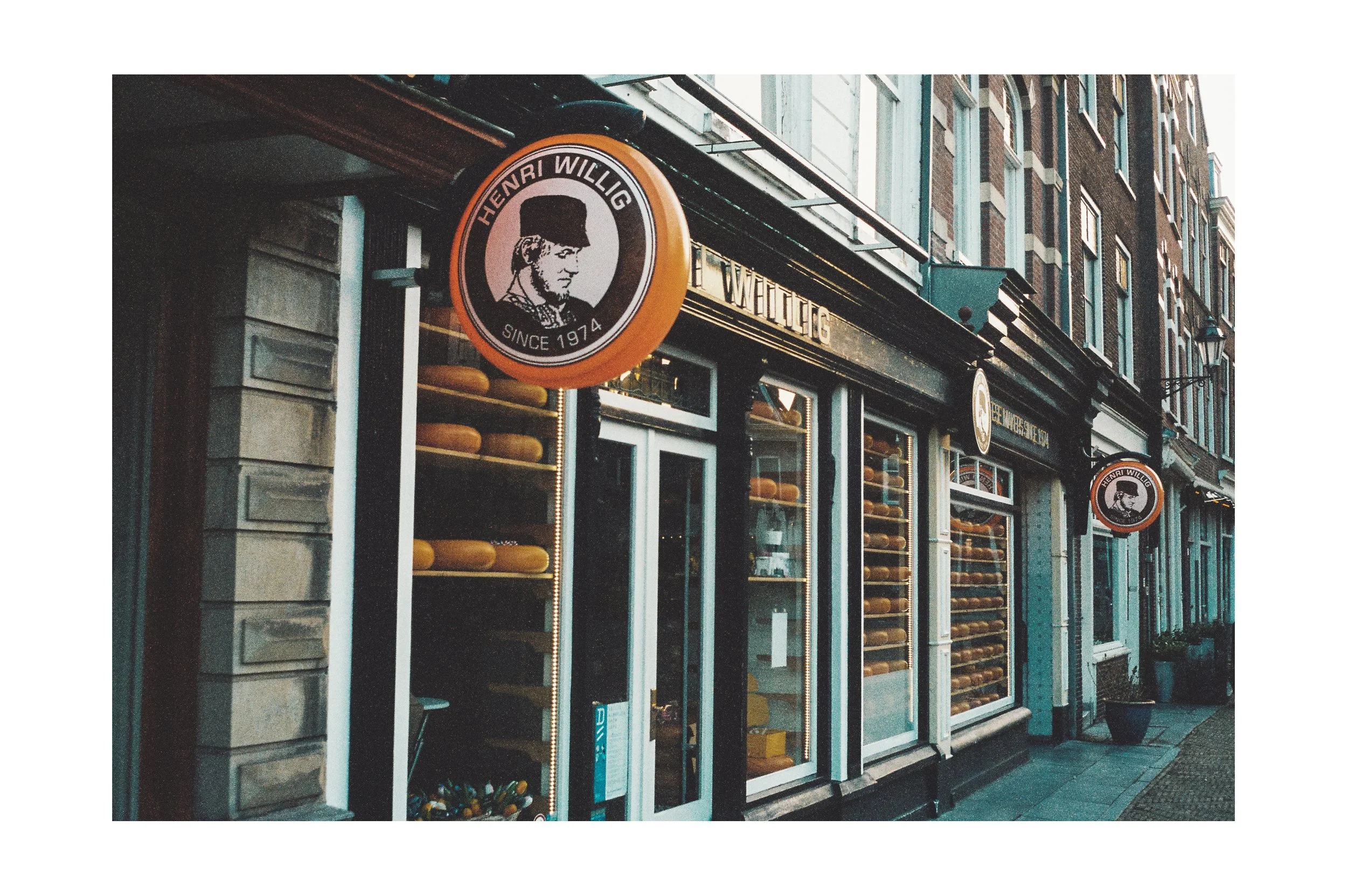 Exterior view of a cheese shop with the sign displaying Henri Willig since 1974, showcasing shelves filled with cheese wheels.