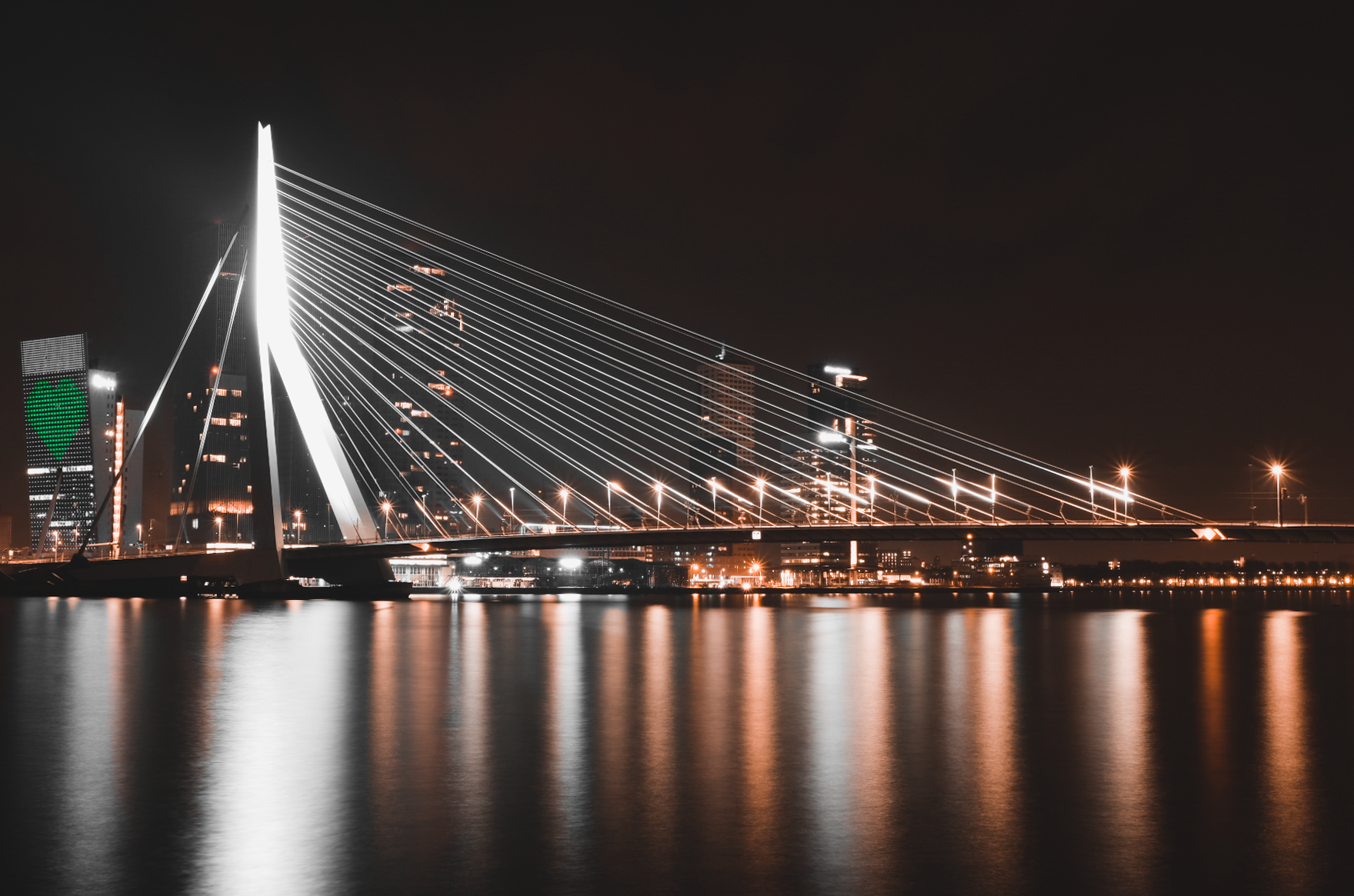 Nighttime cityscape featuring a modern illuminated cable-stayed bridge over a body of water, with city lights reflecting on the water's surface and a skyline with high-rise buildings in the Erasmus Bridge.Netherlands landscape photogtraphy Rotterdam 