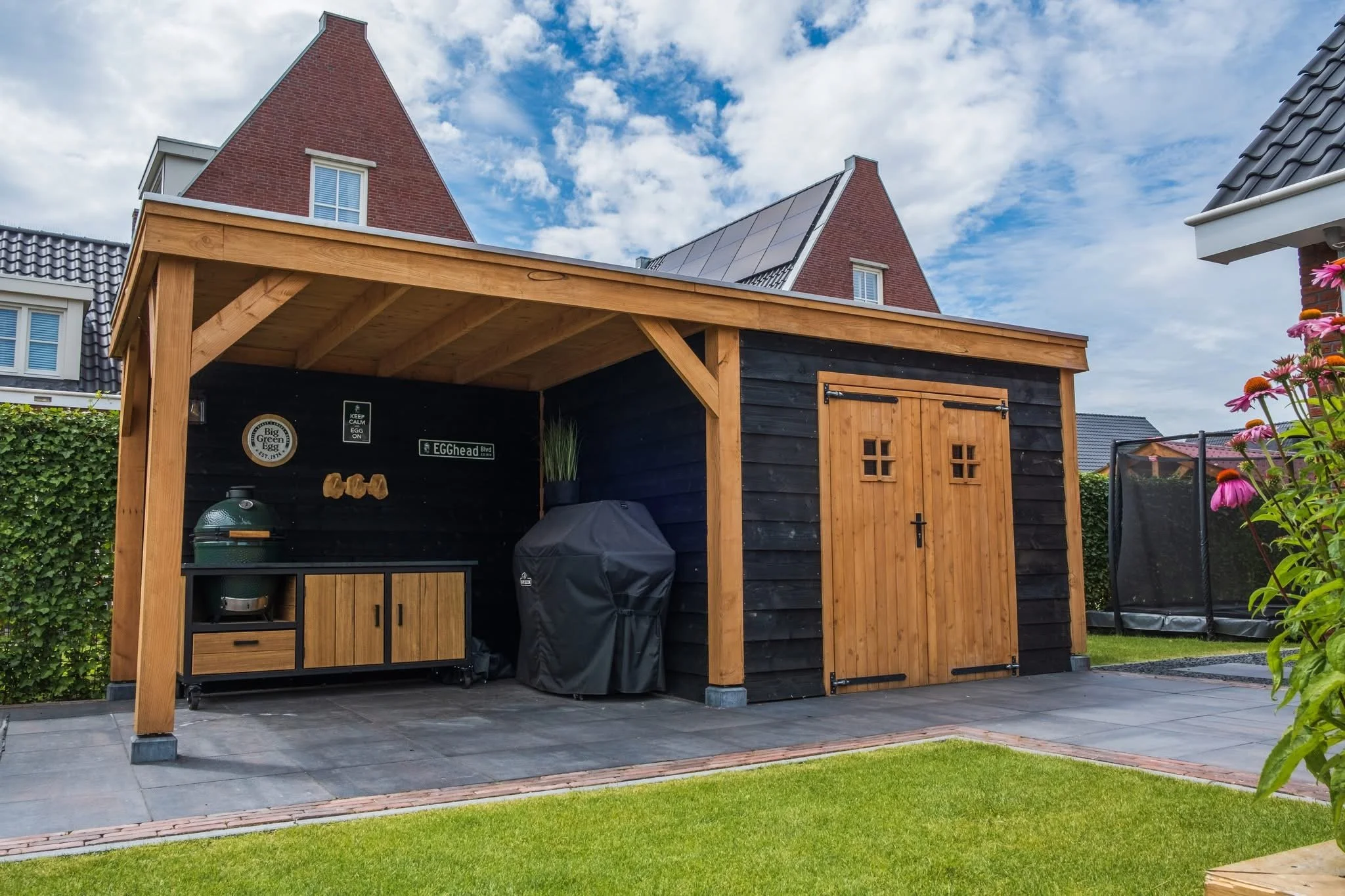 Backyard shed with wooden doors and black walls, surrounded by green grass and garden, with a trampoline in the background.
