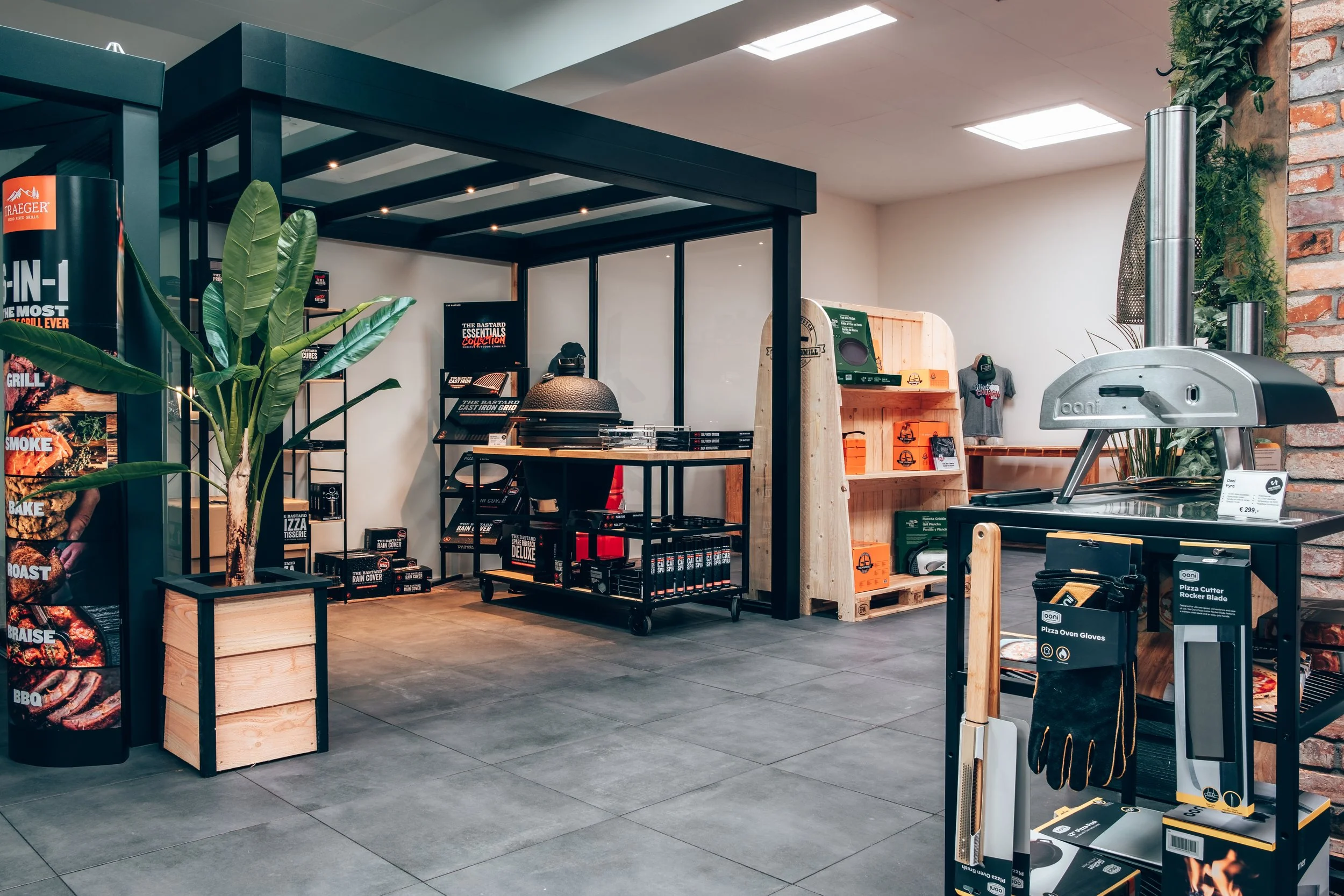 Indoor retail space selling outdoor and barbecue equipment, featuring a large potted plant, a brick wall, and various grilling accessories and tools on display.