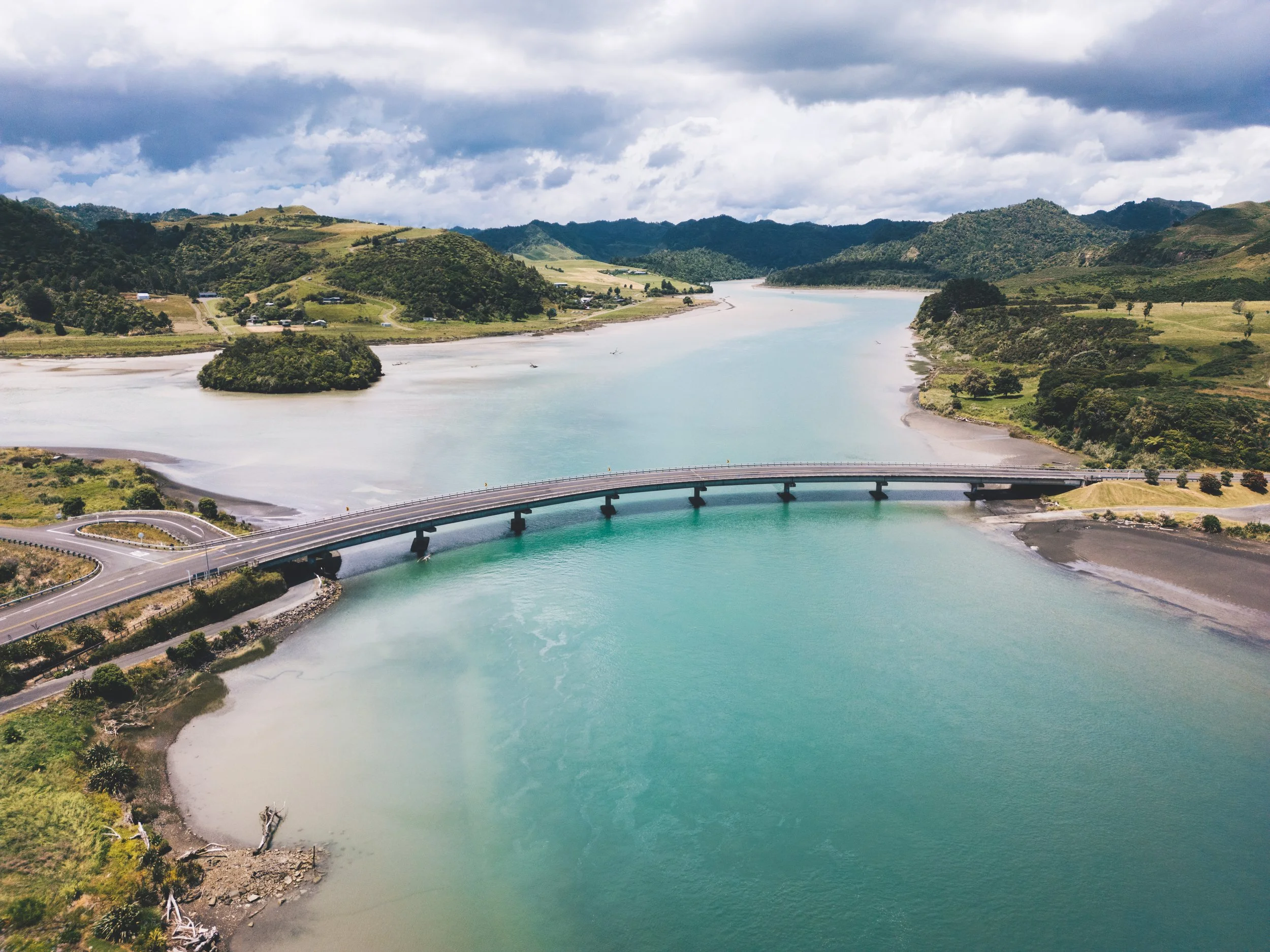 An aerial view of a river with a bridge crossing over it, surrounded by green hills and fields under a partly cloudy sky.