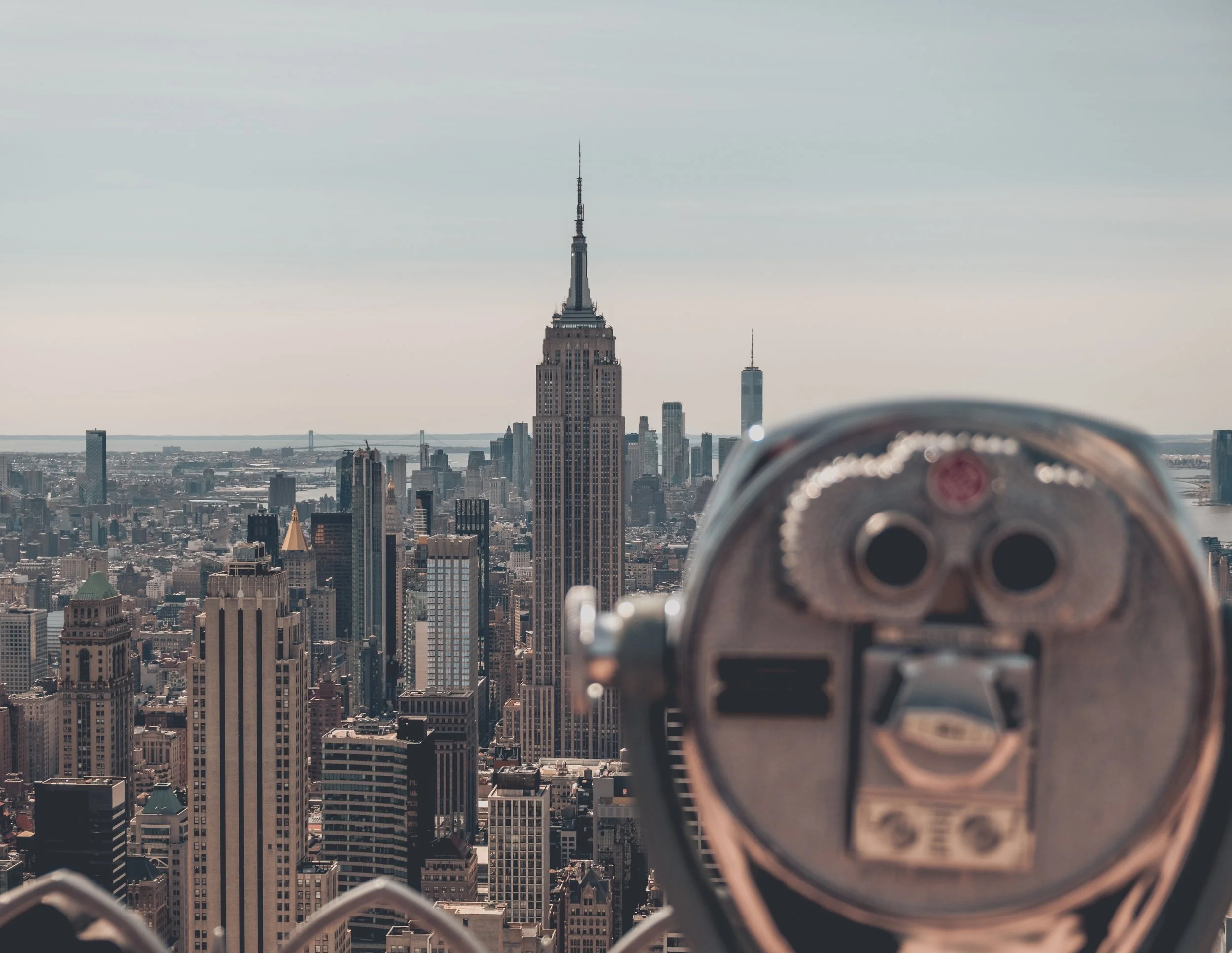 View of New York City skyline from an observation deck with a coin-operated binocular viewer in the foreground, featuring the Empire State Building and One World Trade Center.