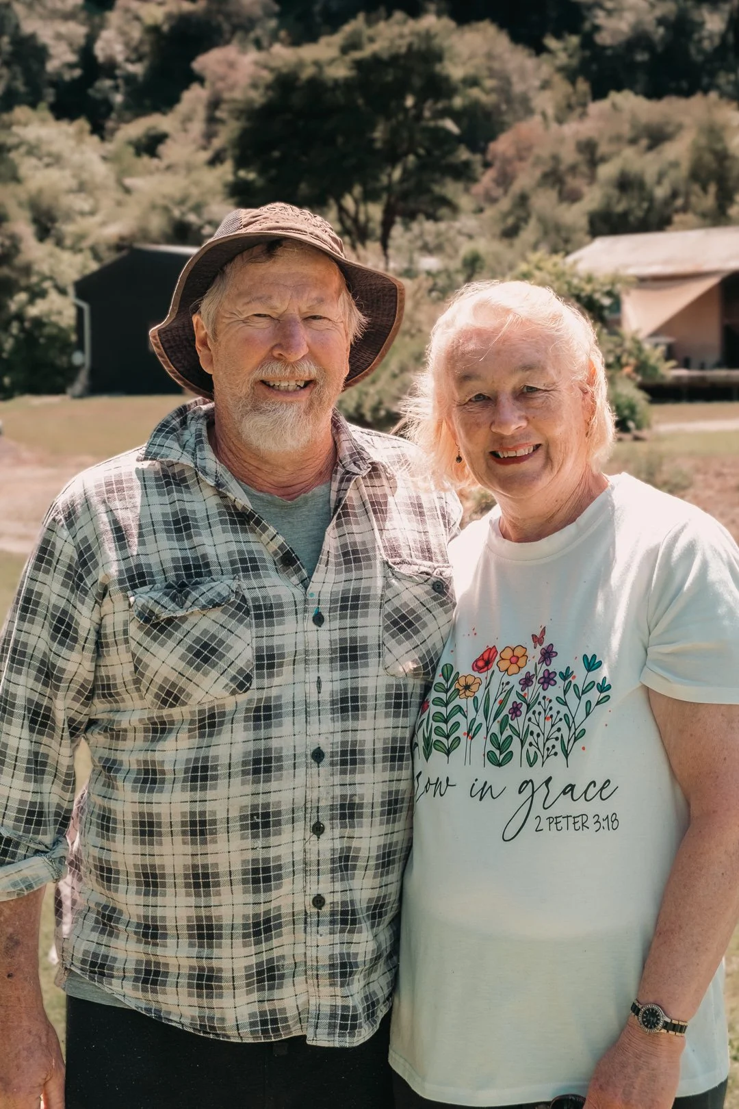 An older man and woman smiling outdoors with trees and buildings in the background.