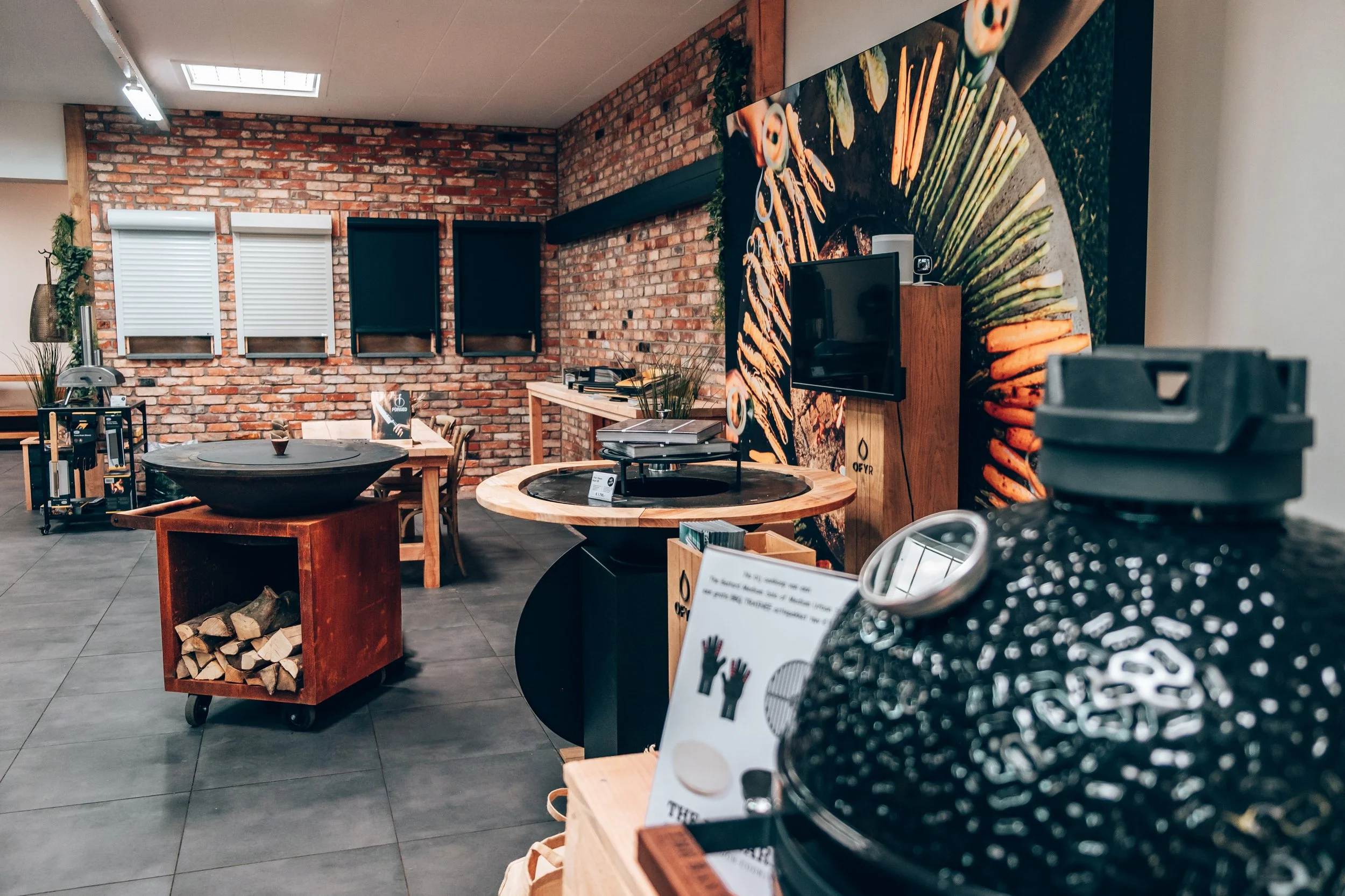 Interior of a store or showroom with brick walls, displaying various kitchen and barbecue accessories, wood logs, and decorative items.