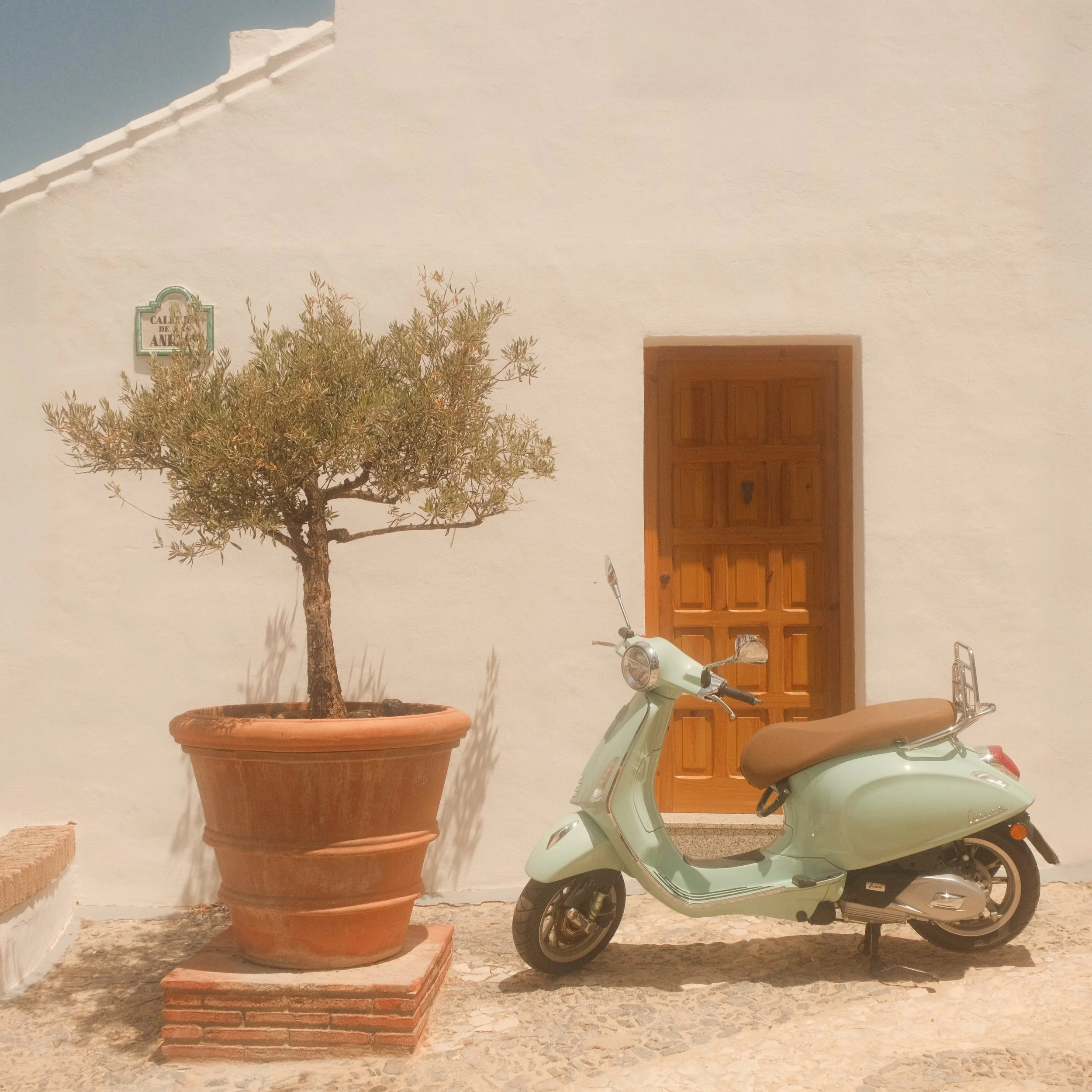 A mint green scooter parked on a stone pavement against a white wall with a wooden door. There is a large terracotta pot with a small tree next to the scooter. A street sign reading 'Calle de' is partially visible on the wall.