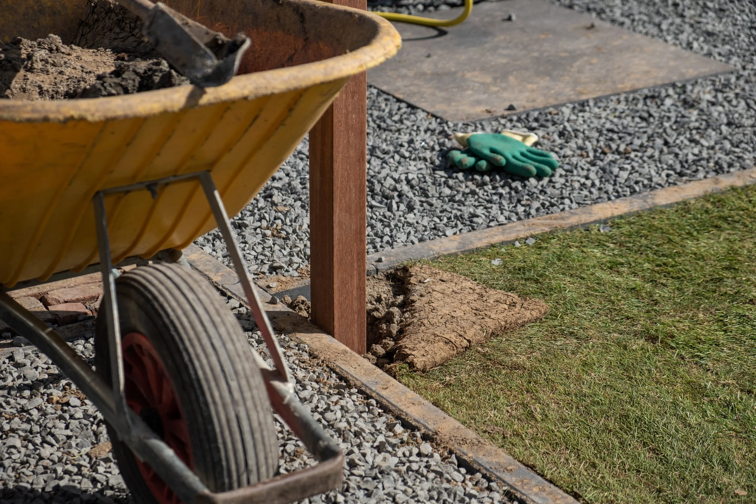 A yellow wheelbarrow filled with dirt, next to a wooden post, with gardening gloves on gravel and grass underneath.