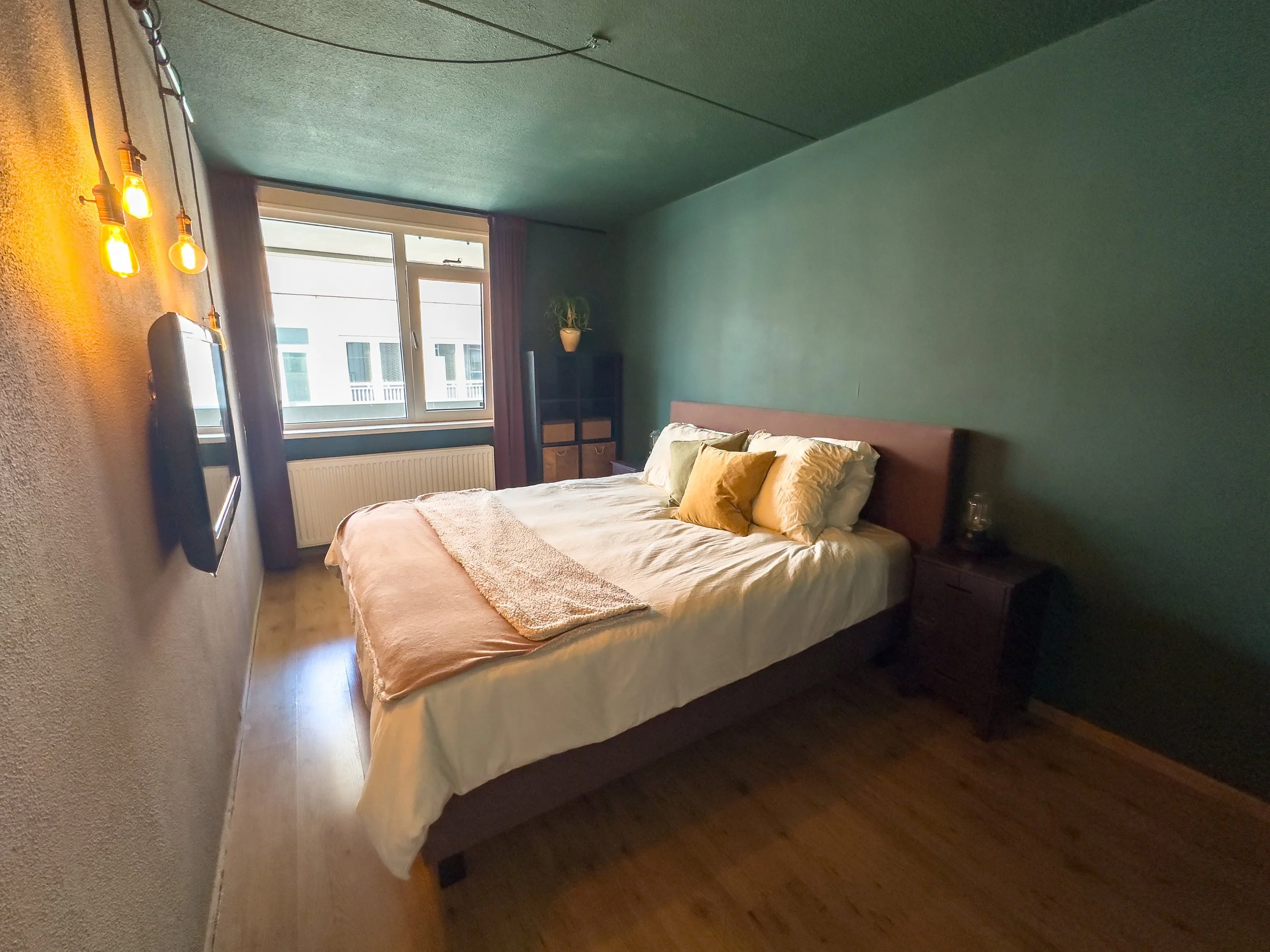 A bedroom with a bed, pillows, wooden nightstand, window, and a wall-mounted TV, with a green and beige color scheme and warm lighting.