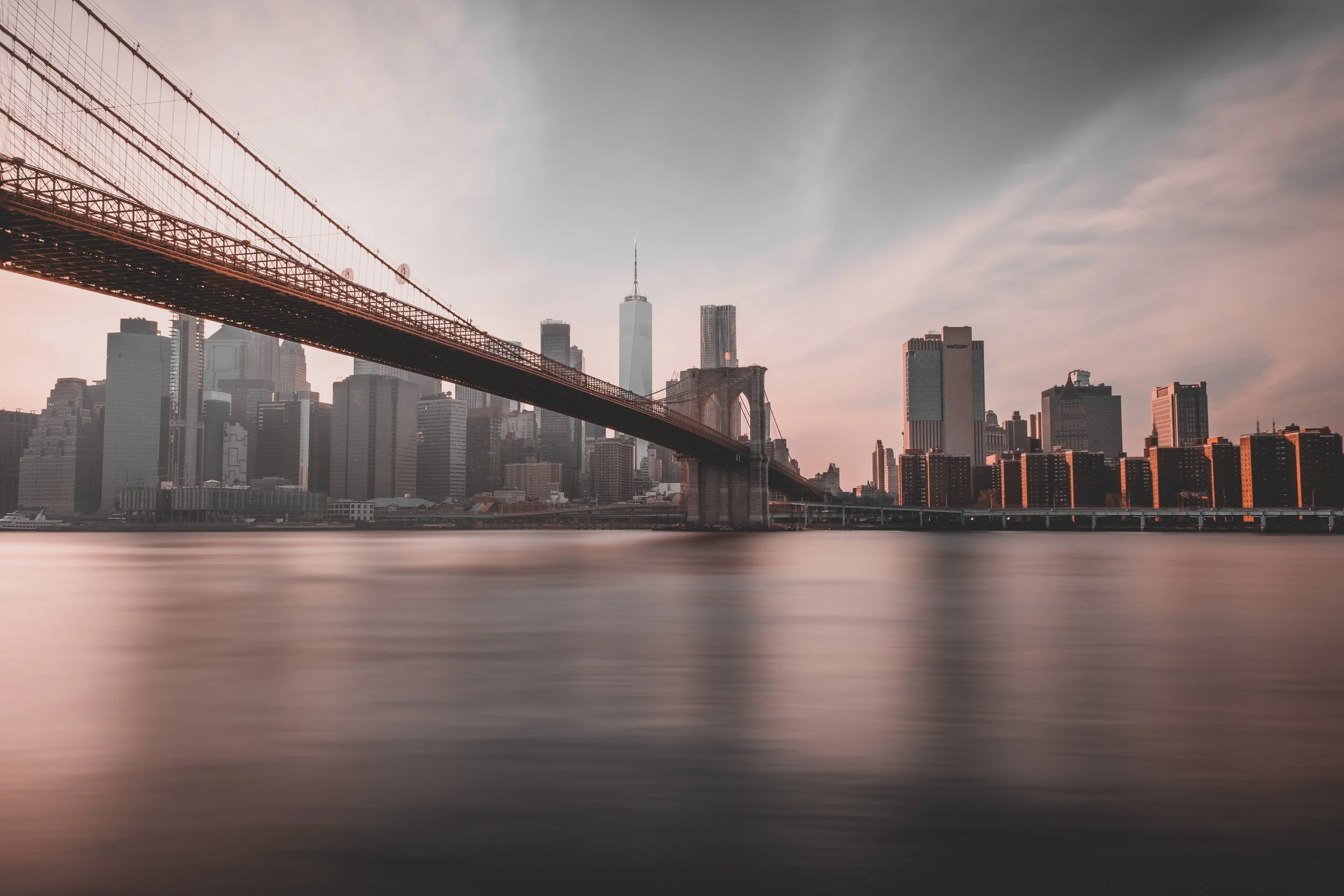 City skyline of New York City with the Brooklyn Bridge in the foreground, water reflection, and skyscrapers including One World Trade Center at sunset.