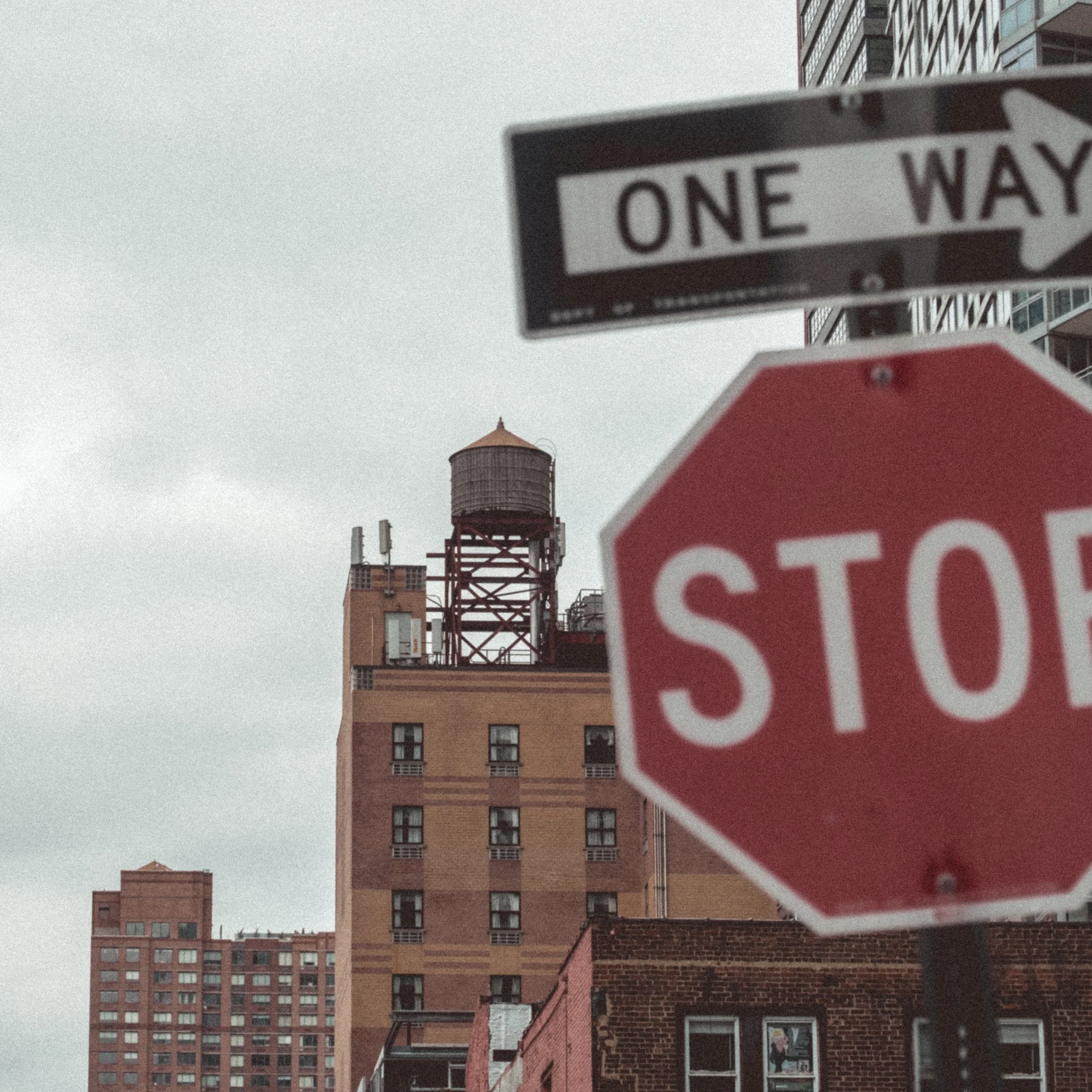 Street corner with stop sign, one-way street sign, high-rise buildings, and a water tower against a cloudy sky.