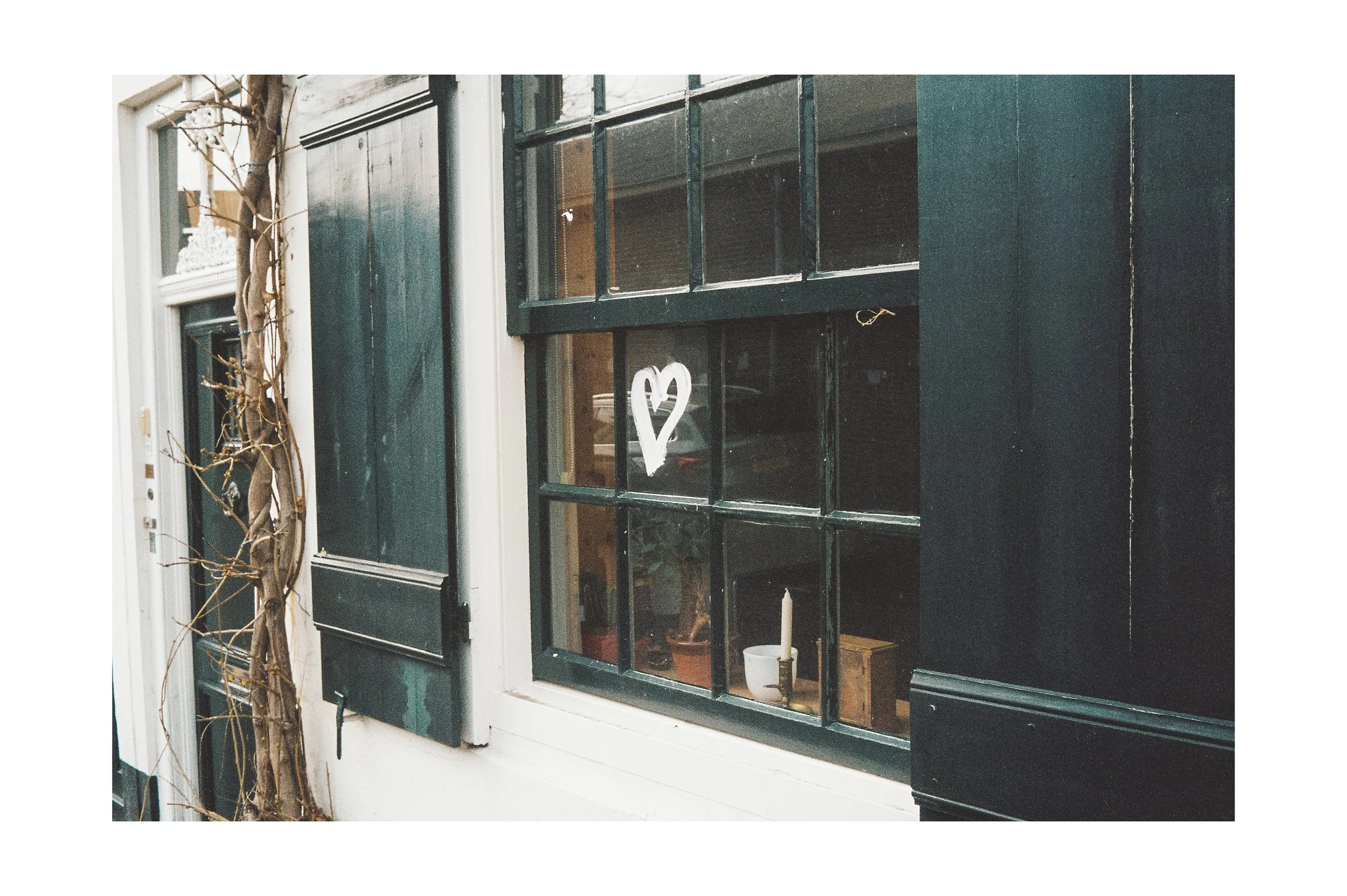Close-up of a black-framed window with a white heart drawn on the glass, next to a vine-covered wall and a candle inside.