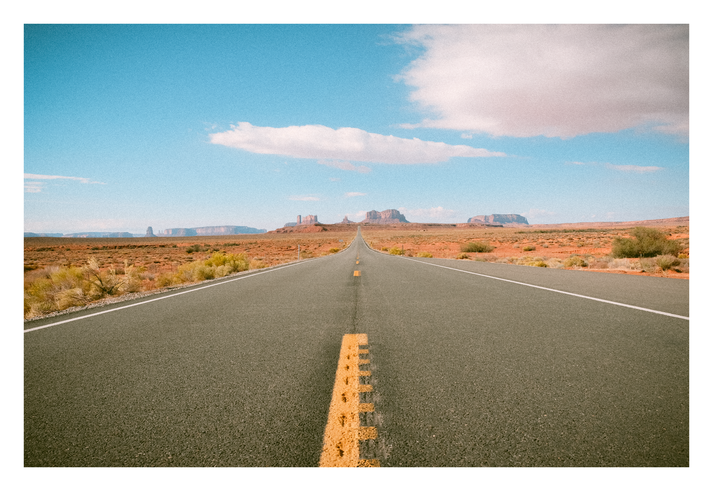 A straight empty desert highway extends into the horizon with reddish rocky formations and mesas in the background under a partly cloudy sky.