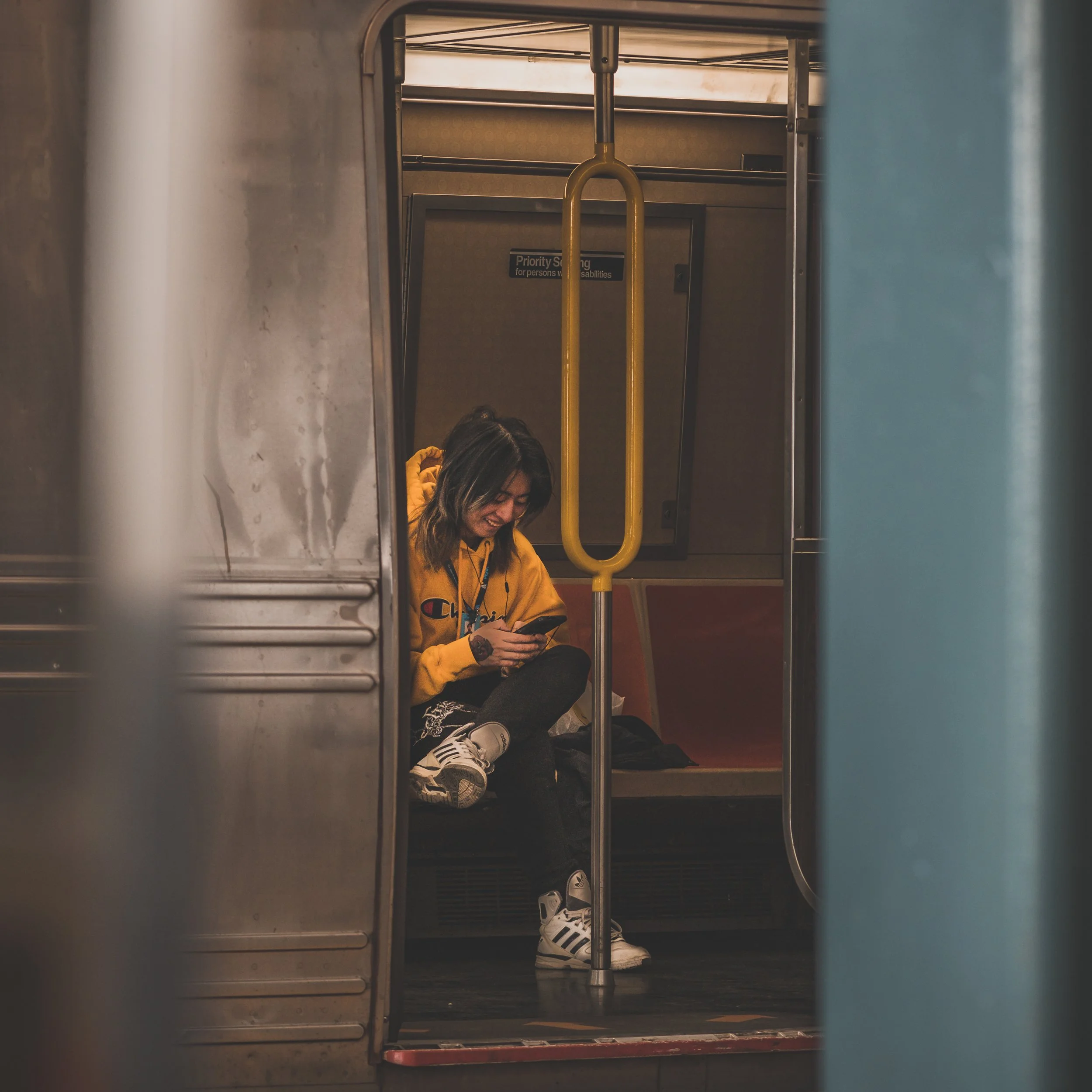 A young woman sitting on a subway train seat, looking at her phone, wearing a yellow hoodie, black pants, and white sneakers.