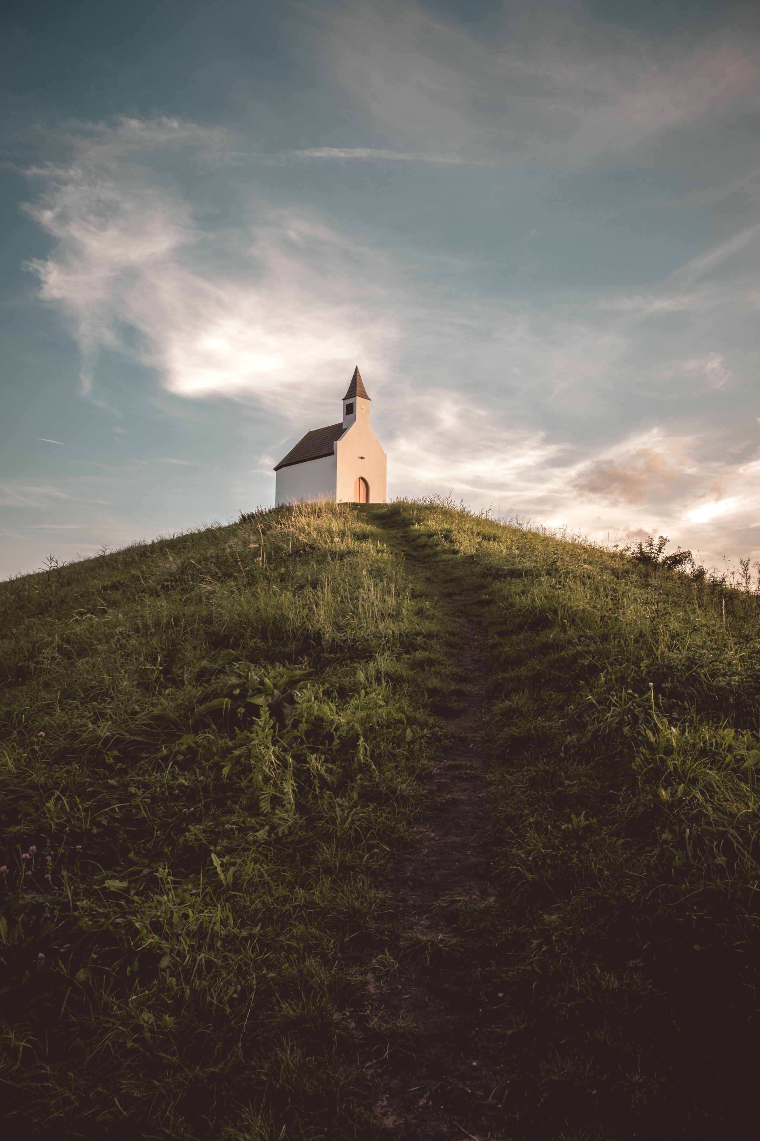 A small white church on top of a grassy hill under a partly cloudy sky at sunset.