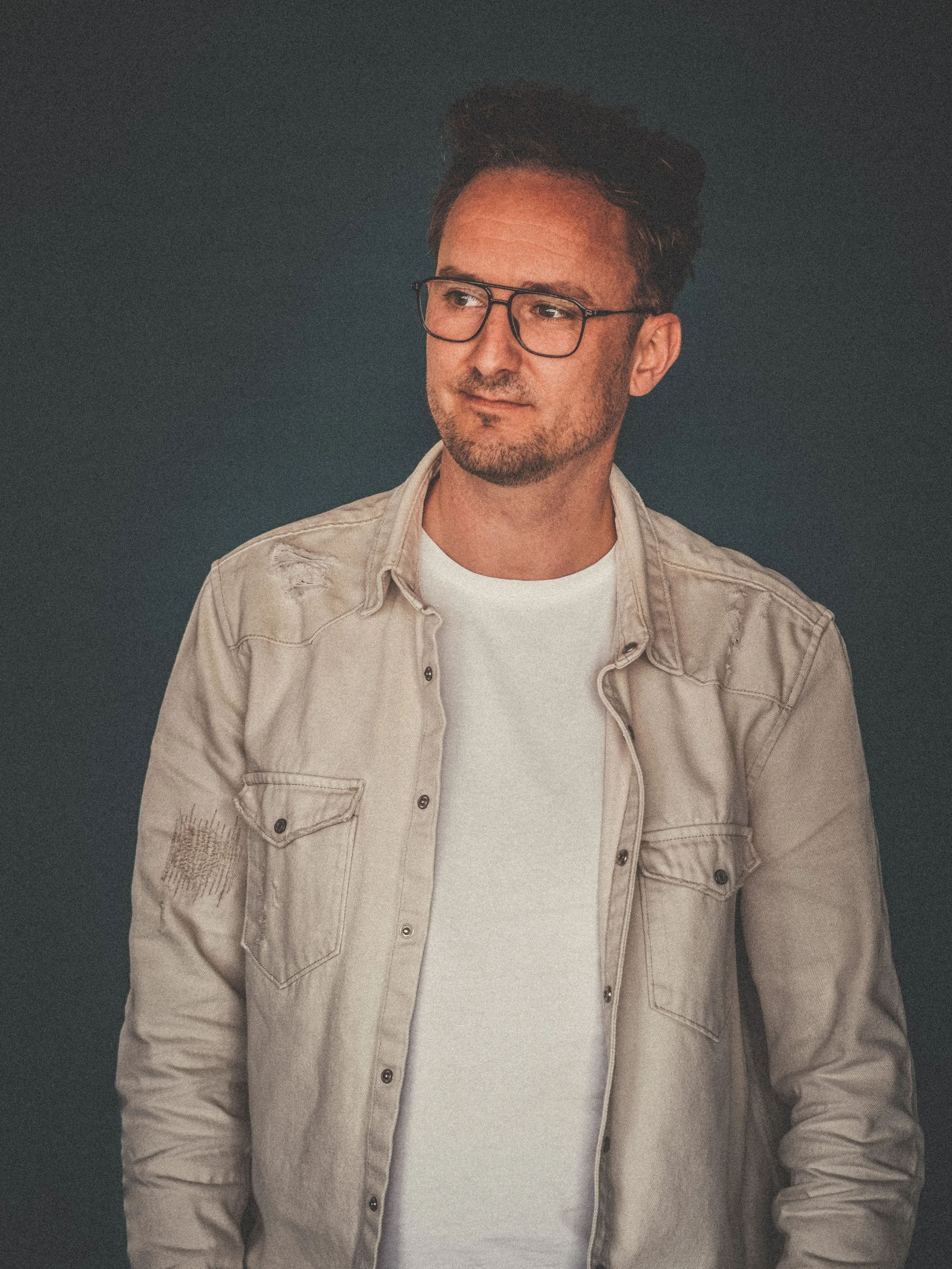 A man with glasses and a beard wearing a beige denim shirt over a white t-shirt, standing against a dark background.