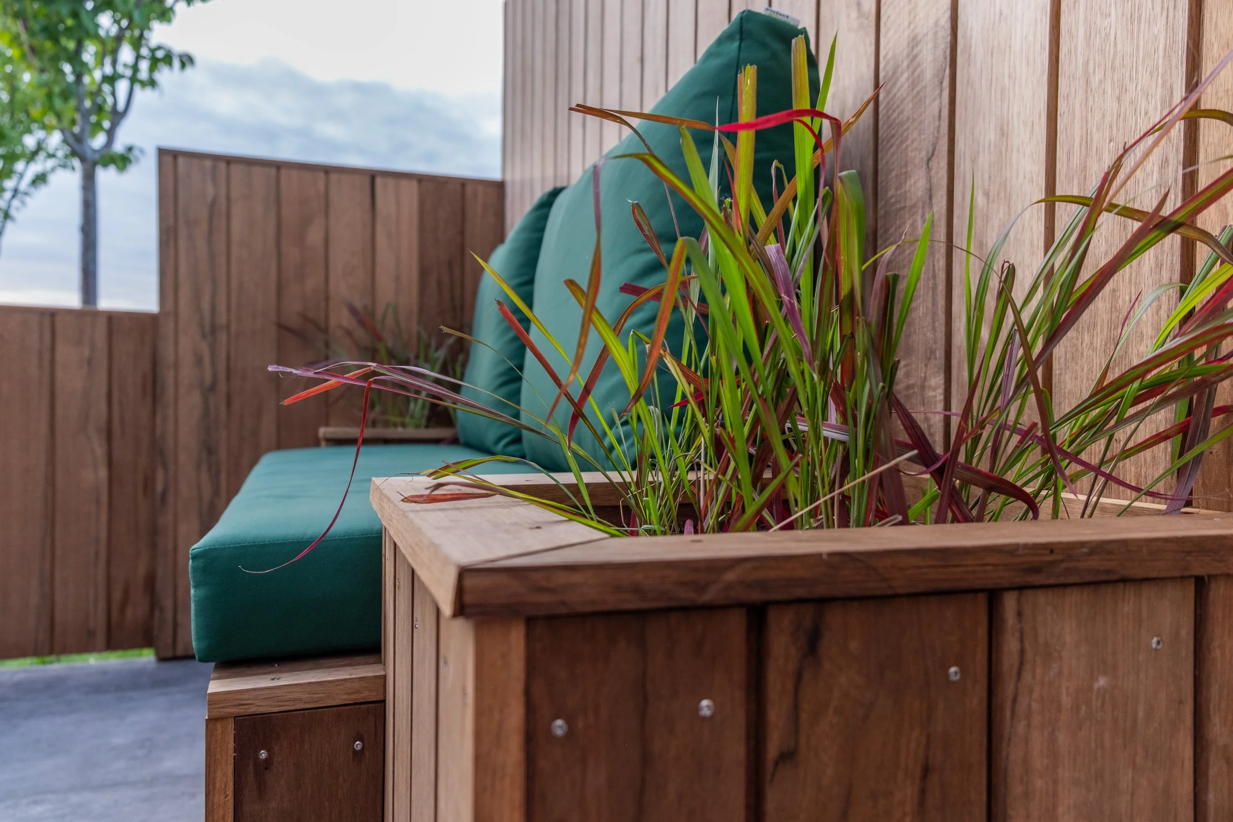 Close-up of a modern outdoor patio with wooden furniture, a teal cushioned bench with pillows, and a planter with green and purple plants against a wood-paneled wall, under a cloudy sky.