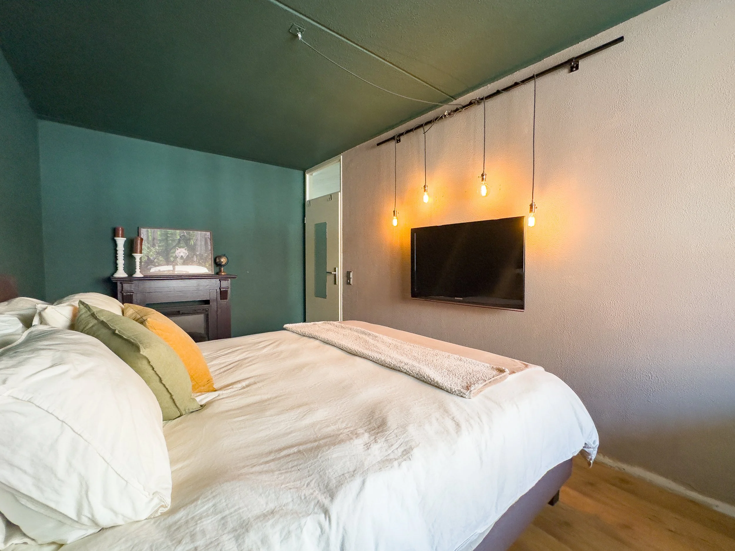 A cozy bedroom with a large bed featuring white linens and multiple pillows, a dark wooden headboard, and wall-mounted string lights with visible bulbs. There's a flat-screen TV on the wall opposite the bed and a small decorative shelf or picture fra
