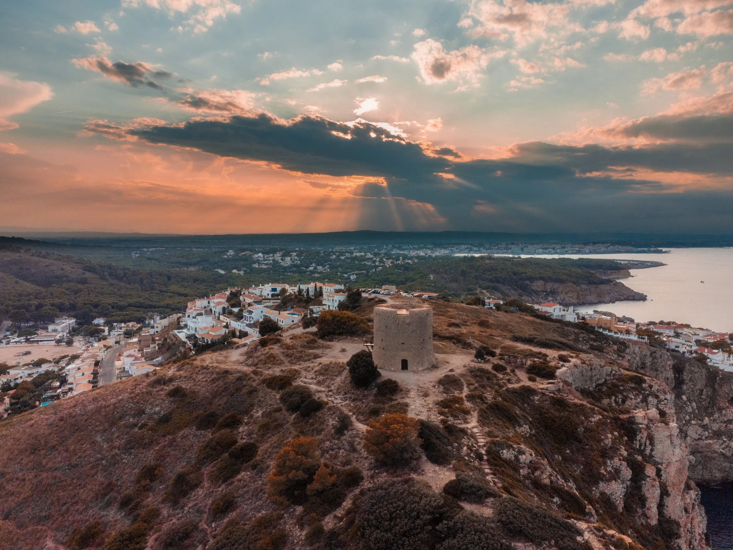 Aerial view of a coastal town with white buildings, a historic tower on a hilltop, ocean in the background, and a dramatic sunset with clouds.