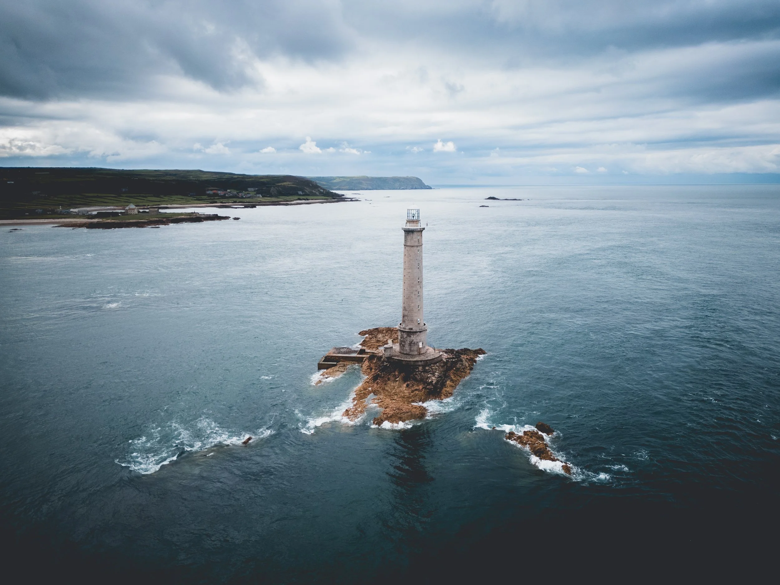 A lighthouse on a rocky island surrounded by the ocean, with a cloudy sky overhead and a distant shoreline visible in the background.