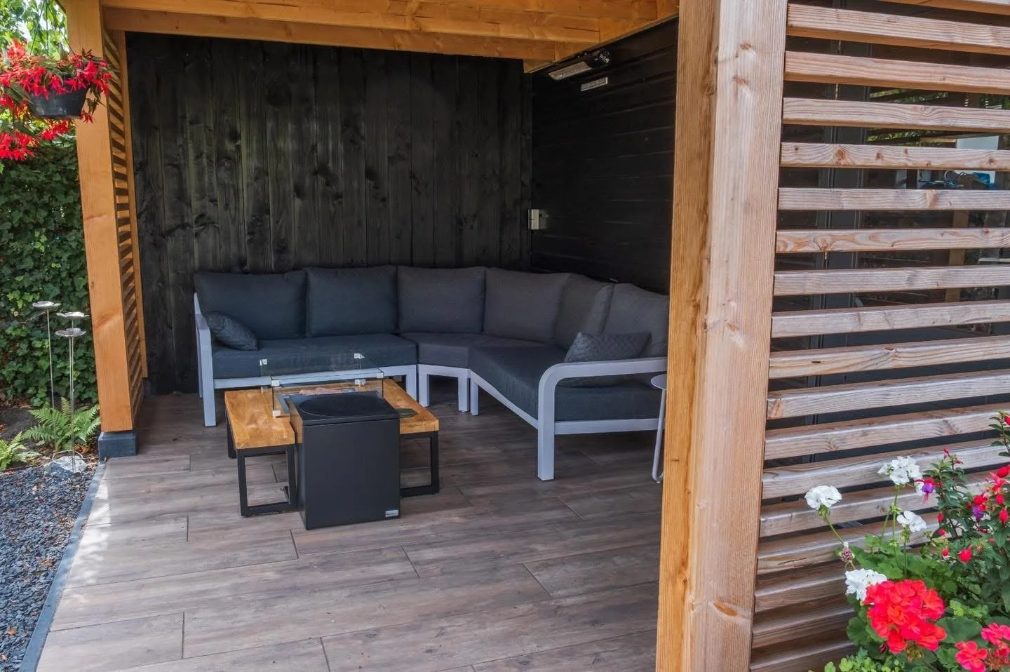 Outdoor wooden gazebo with gray cushioned sectional sofa, small wooden coffee table, and decorative pillows, surrounded by plants and flowers.