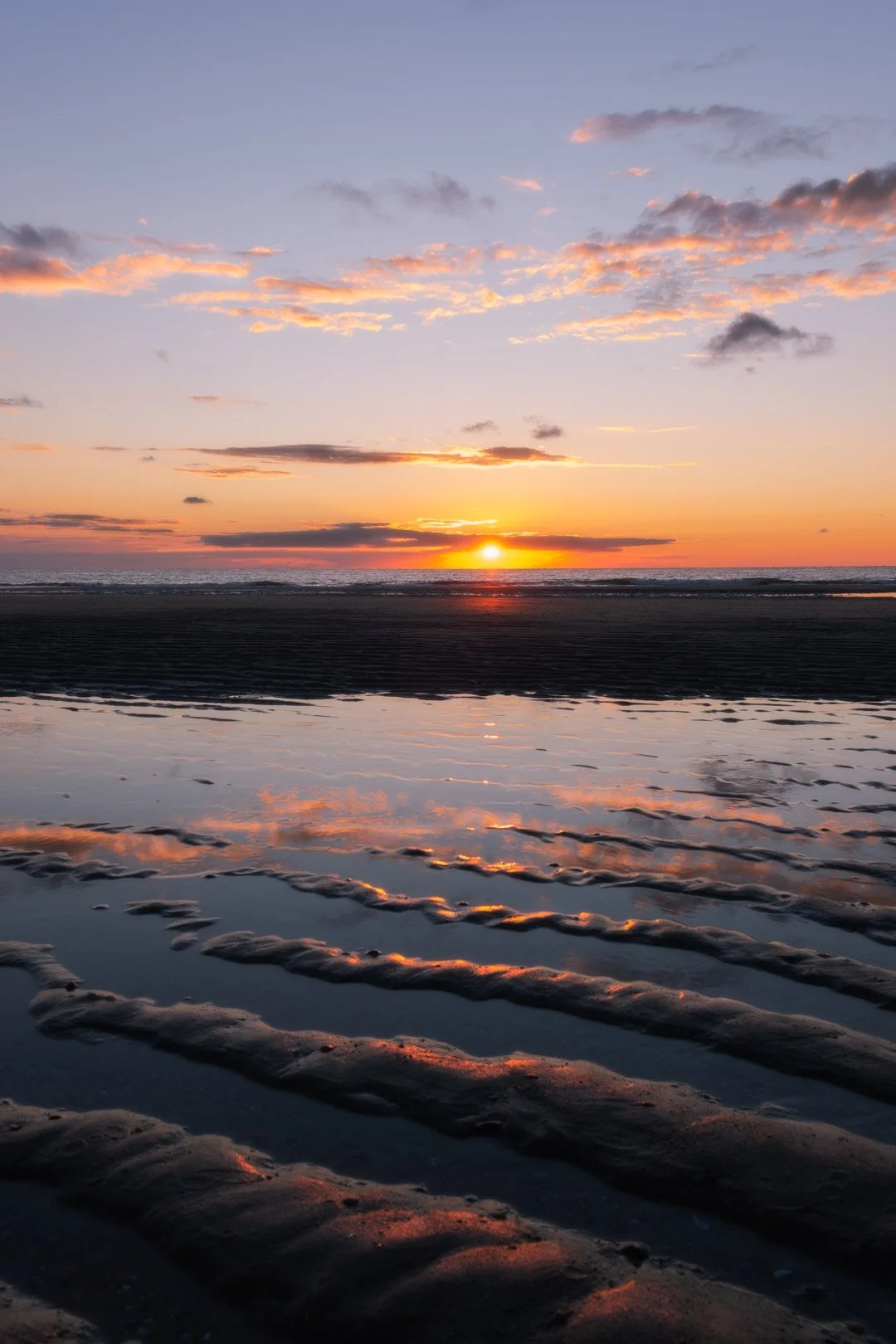 Sunset over the ocean with a sky filled with clouds, and wet sand with rippled reflections in the foreground.