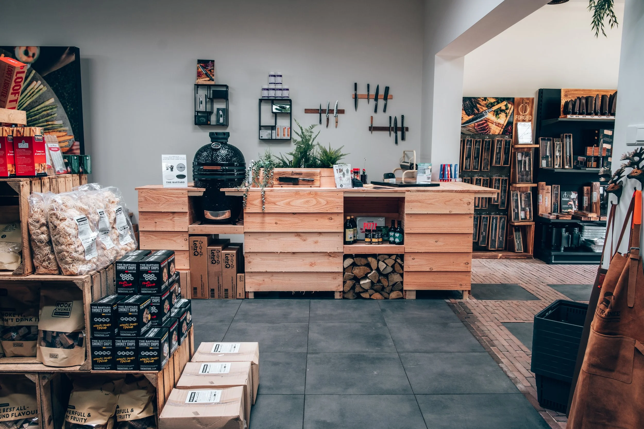 Interior of a store with a wooden counter, shelves with knives, a grill, and firewood. The scene includes snack boxes, bags of chips, and other food items.
