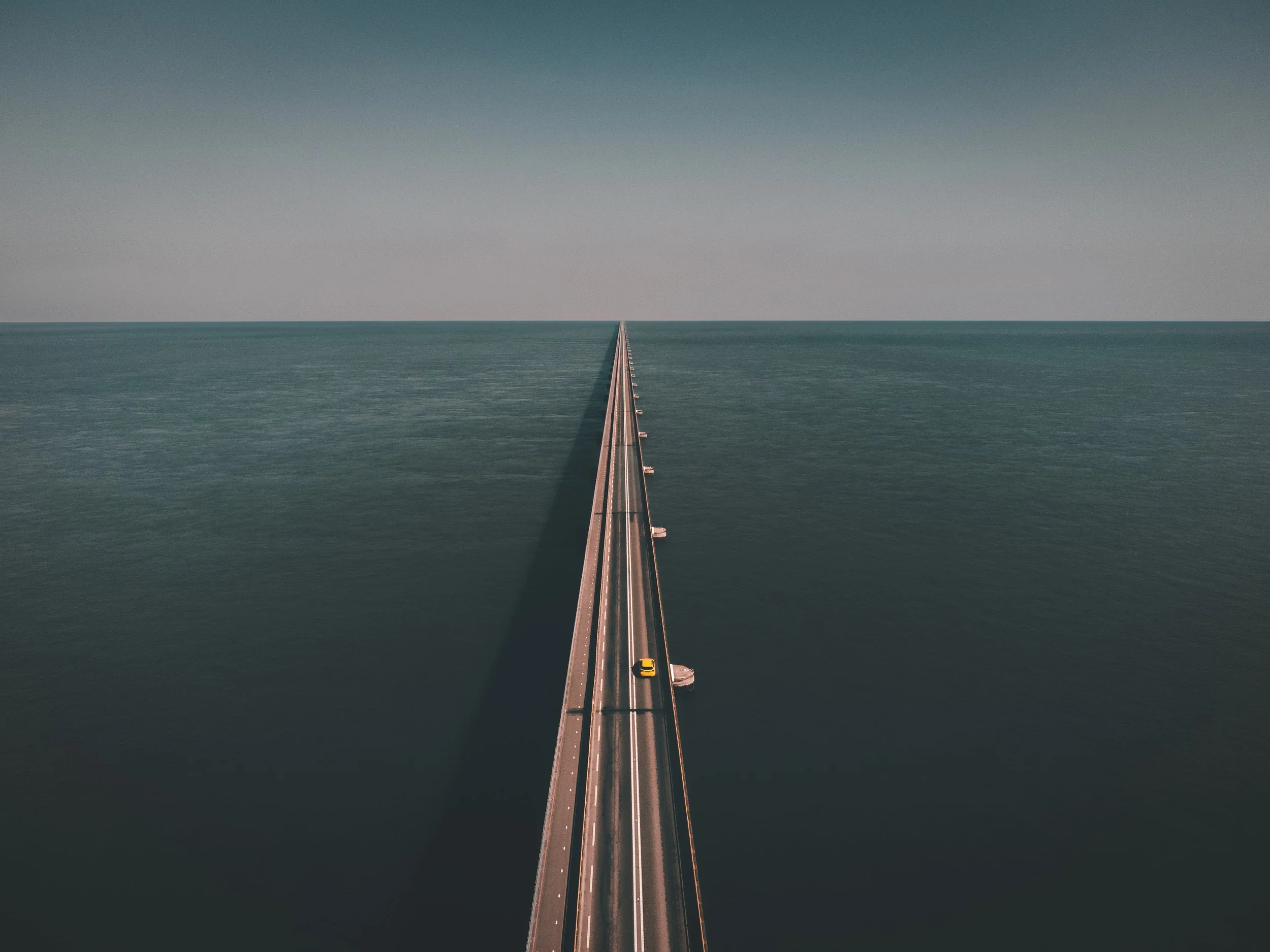 Aerial view of a long, straight bridge extending over a large body of water, with a few cars traveling on it.
