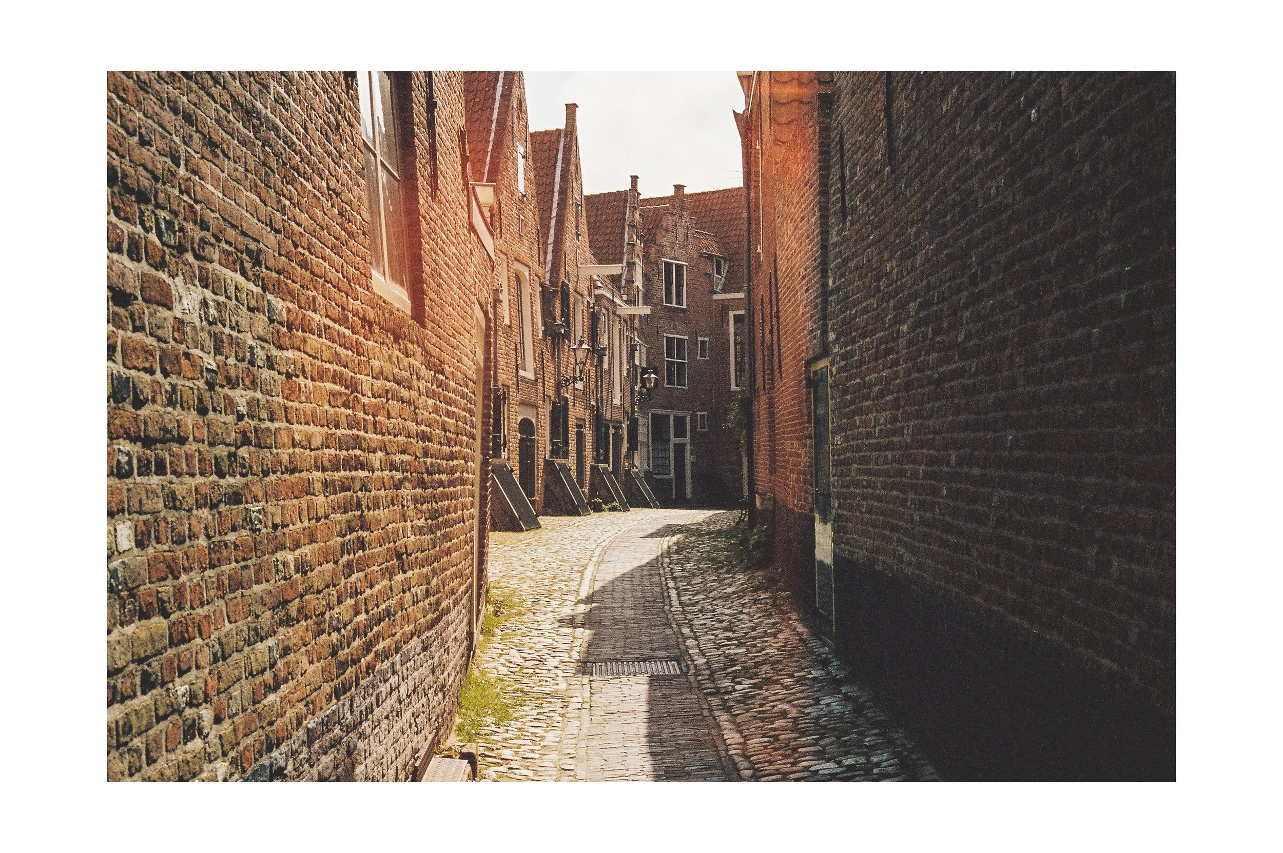 Narrow cobblestone alleyway between brick buildings with small windows and doors, sunlight shining on the street.