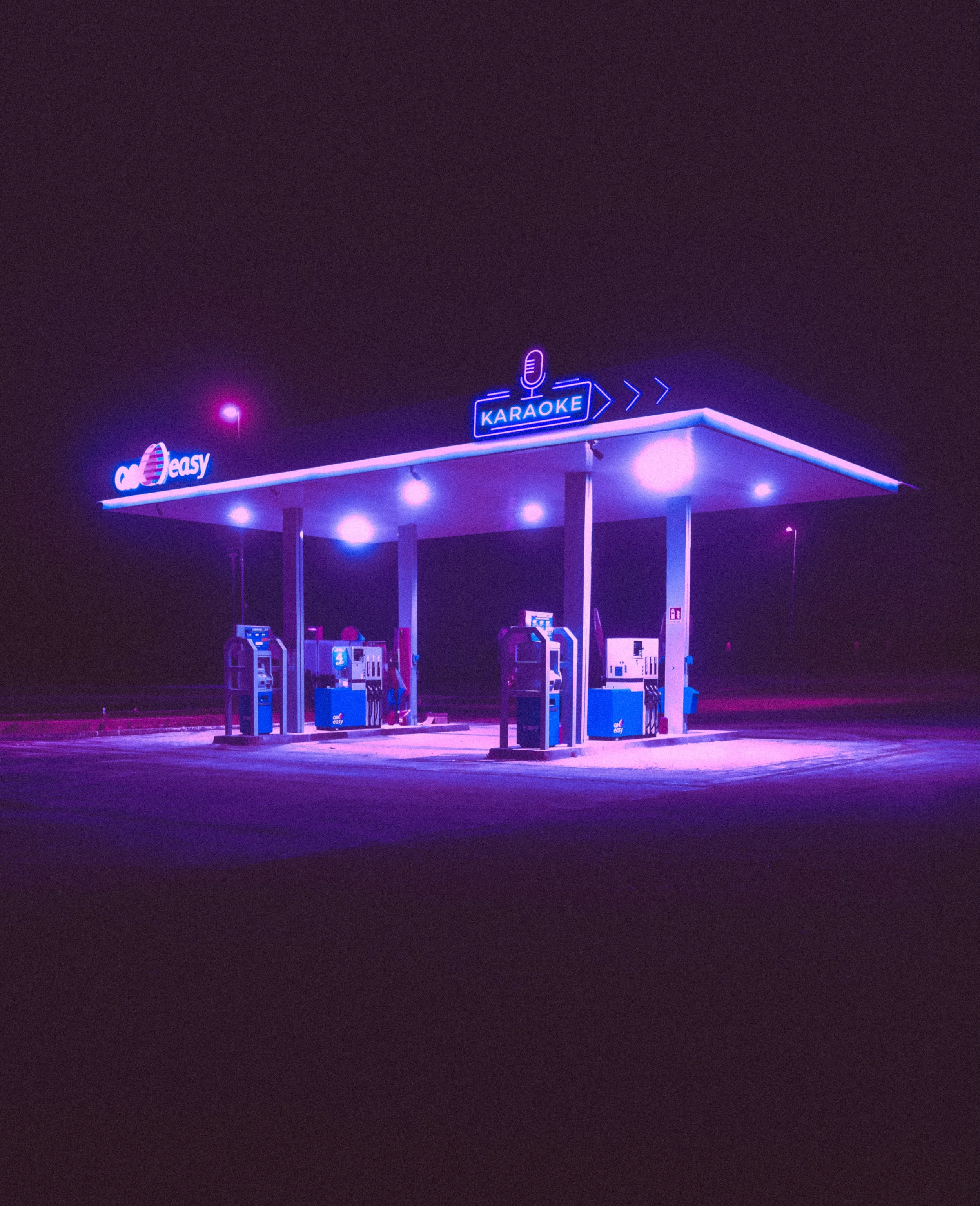 Empty gas station at night illuminated by purple and blue neon lights, with signs reading 'Karaoke' and 'Qeasy'. No cars or people present.