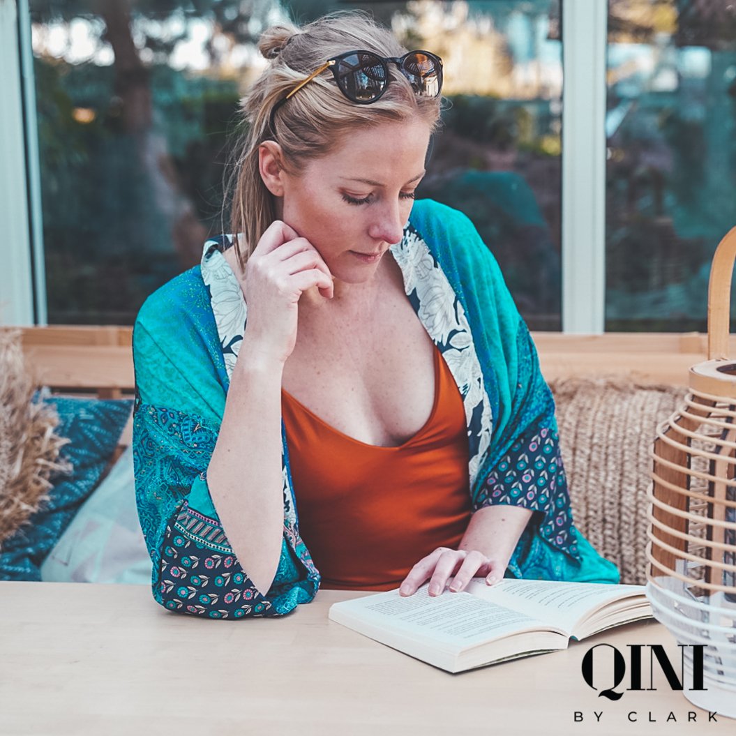 A woman with sunglasses on her head reading a book at a table, with wicker decor and a window behind her.