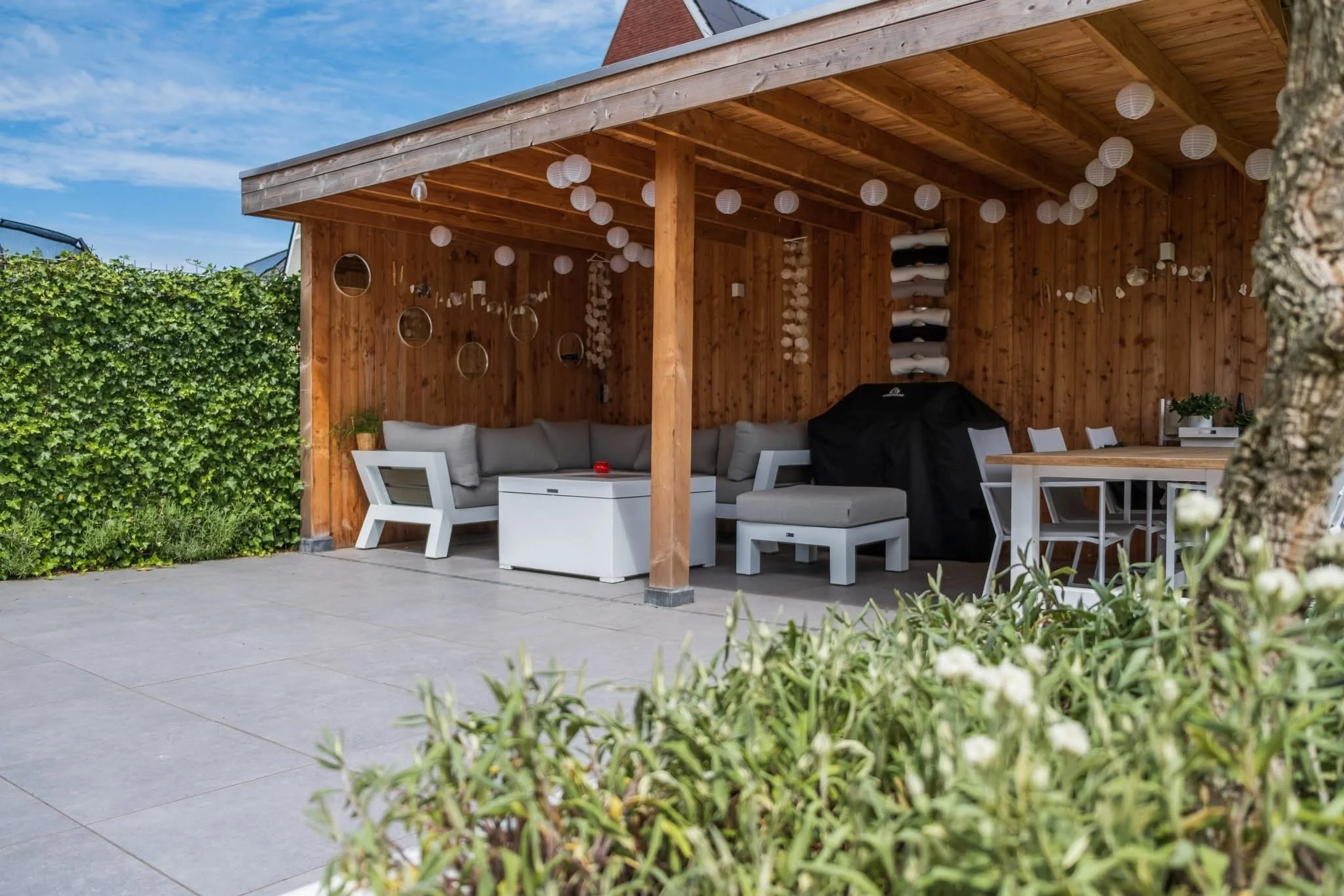 Outdoor patio with a wooden-covered seating area, string lights, white outdoor furniture, a grill covered with a black cover, and decorative hanging elements on the wooden walls. There is greenery in the foreground and a hedge on the left side.