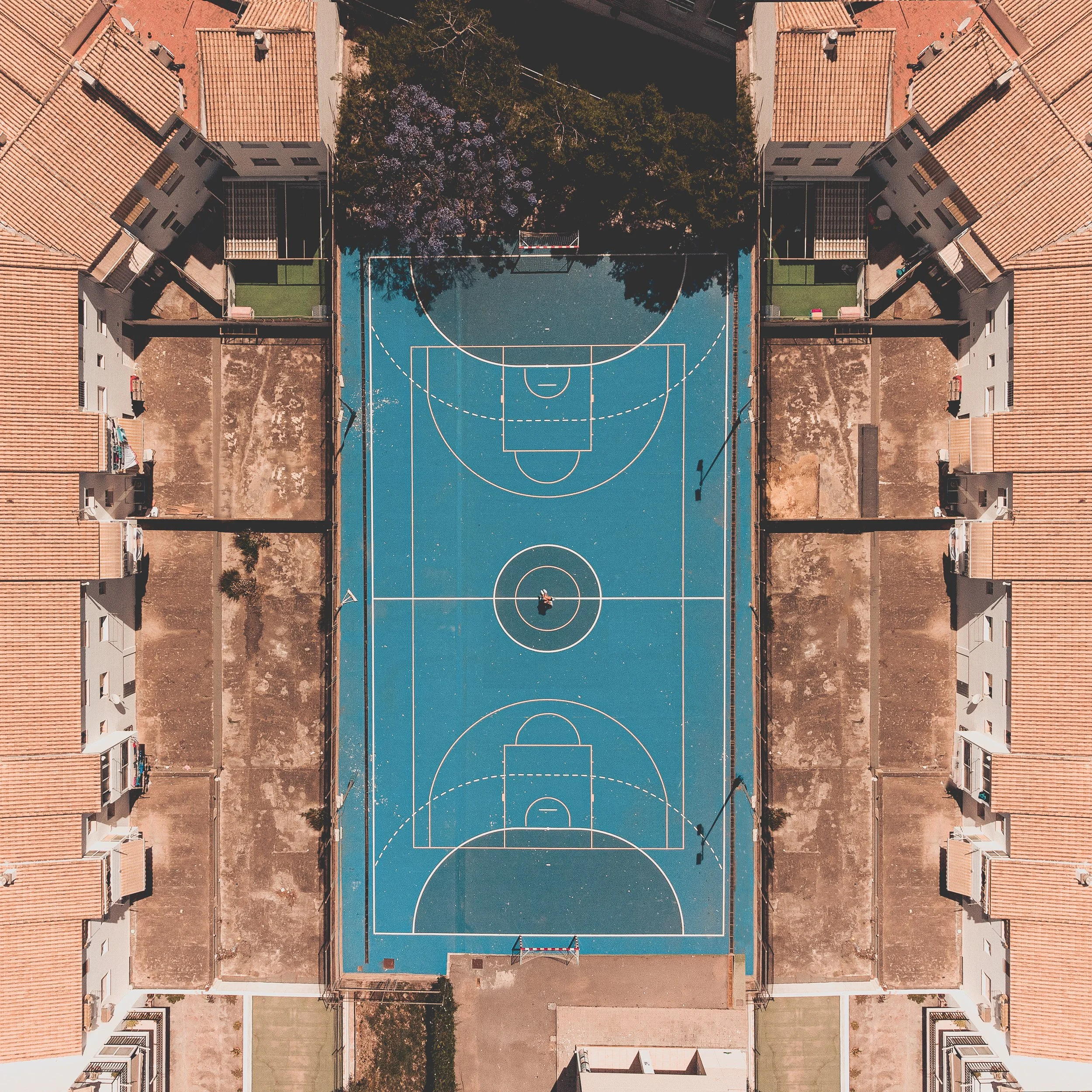 An aerial view of a blue basketball court surrounded by residential buildings, with a person standing in the center of the court and trees near the top of the image.