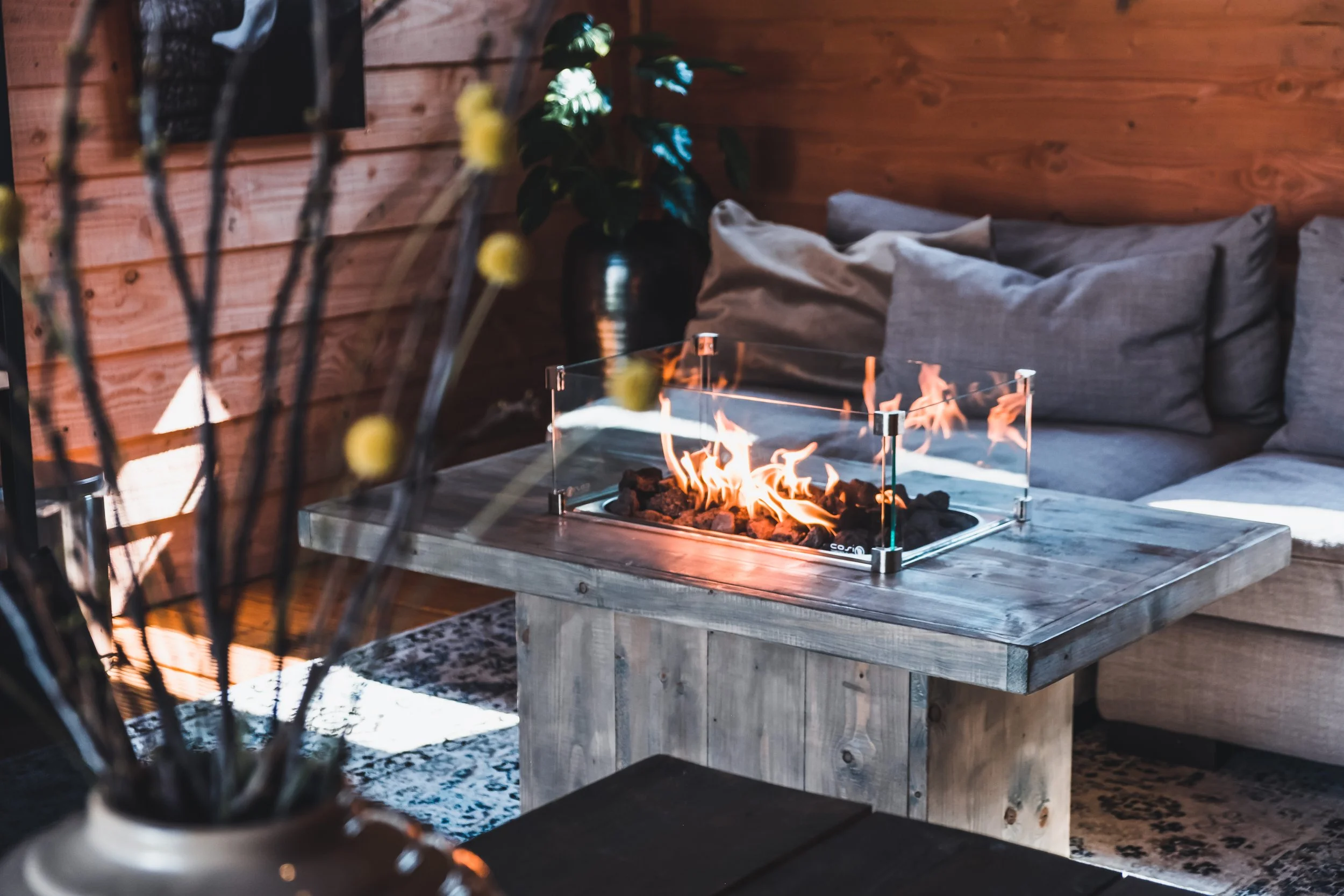 Indoor living space with a glass-encased fireplace on a rustic wooden table. Couch with gray pillows is in the background, and a potted plant with green leaves is next to a wood-paneled wall.