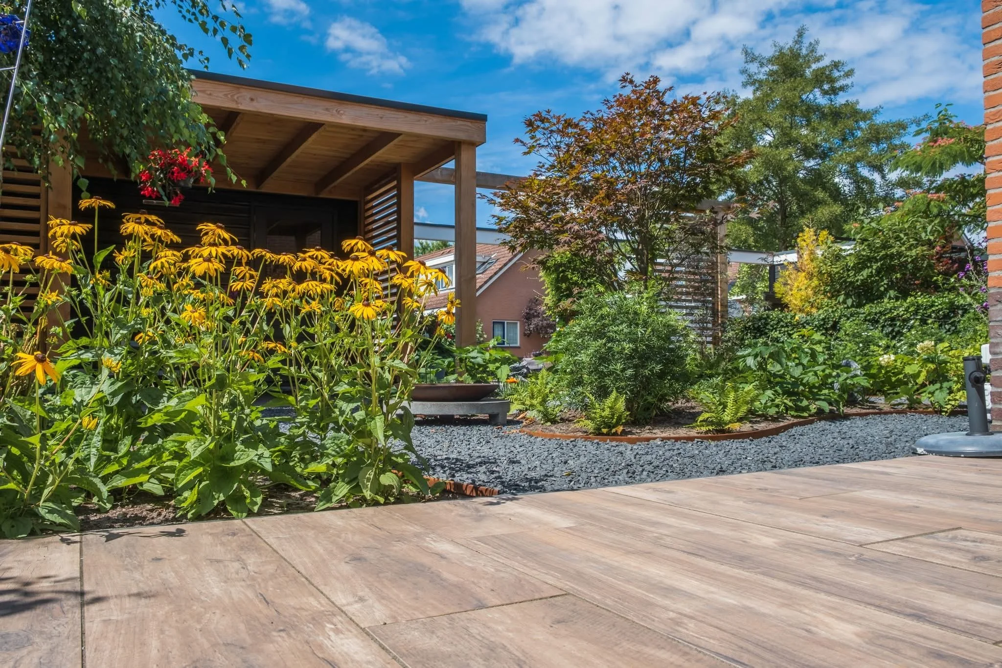 A backyard garden with a wooden deck, bright yellow flowers, green shrubs, trees, and a covered patio area under a blue sky with some clouds.