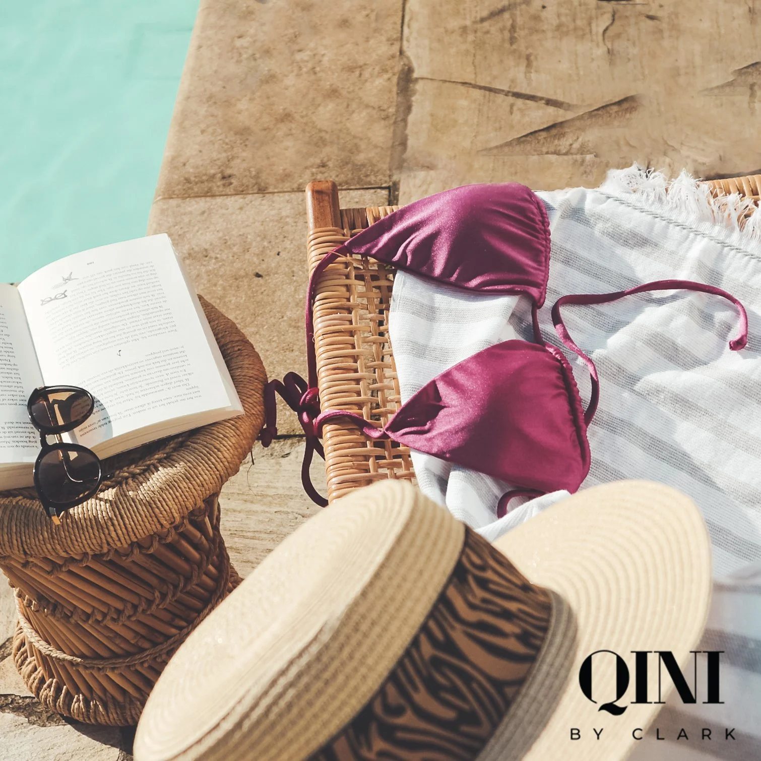 A wicker table with sunglasses, an open book, and a large straw hat next to a pool. A white and gray striped towel is spread out with a pink bikini top on top of it. The pool's water can be seen in the background.