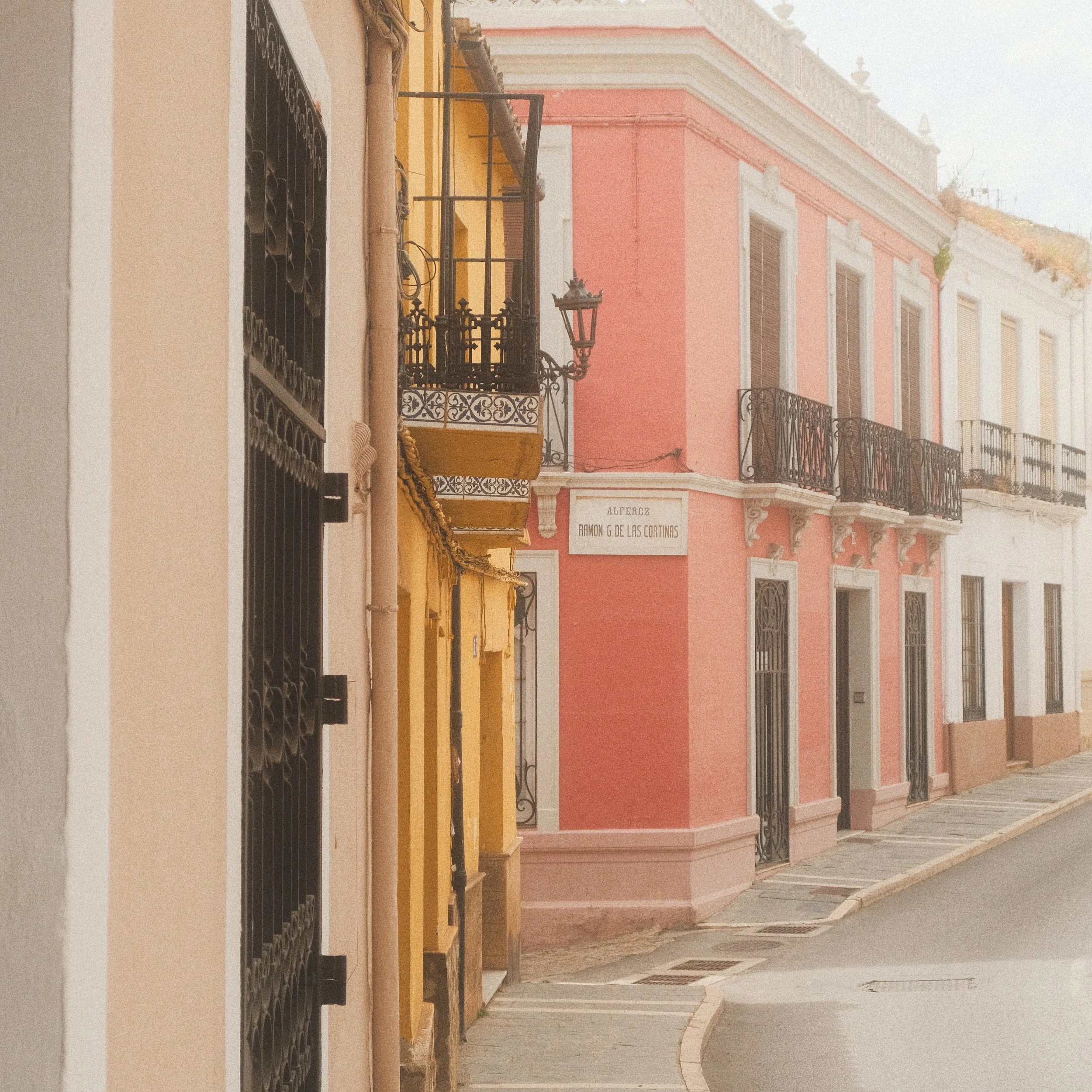 Colorful buildings on a narrow street, including a pink building with black balconies and a yellow building with decorative ironwork.