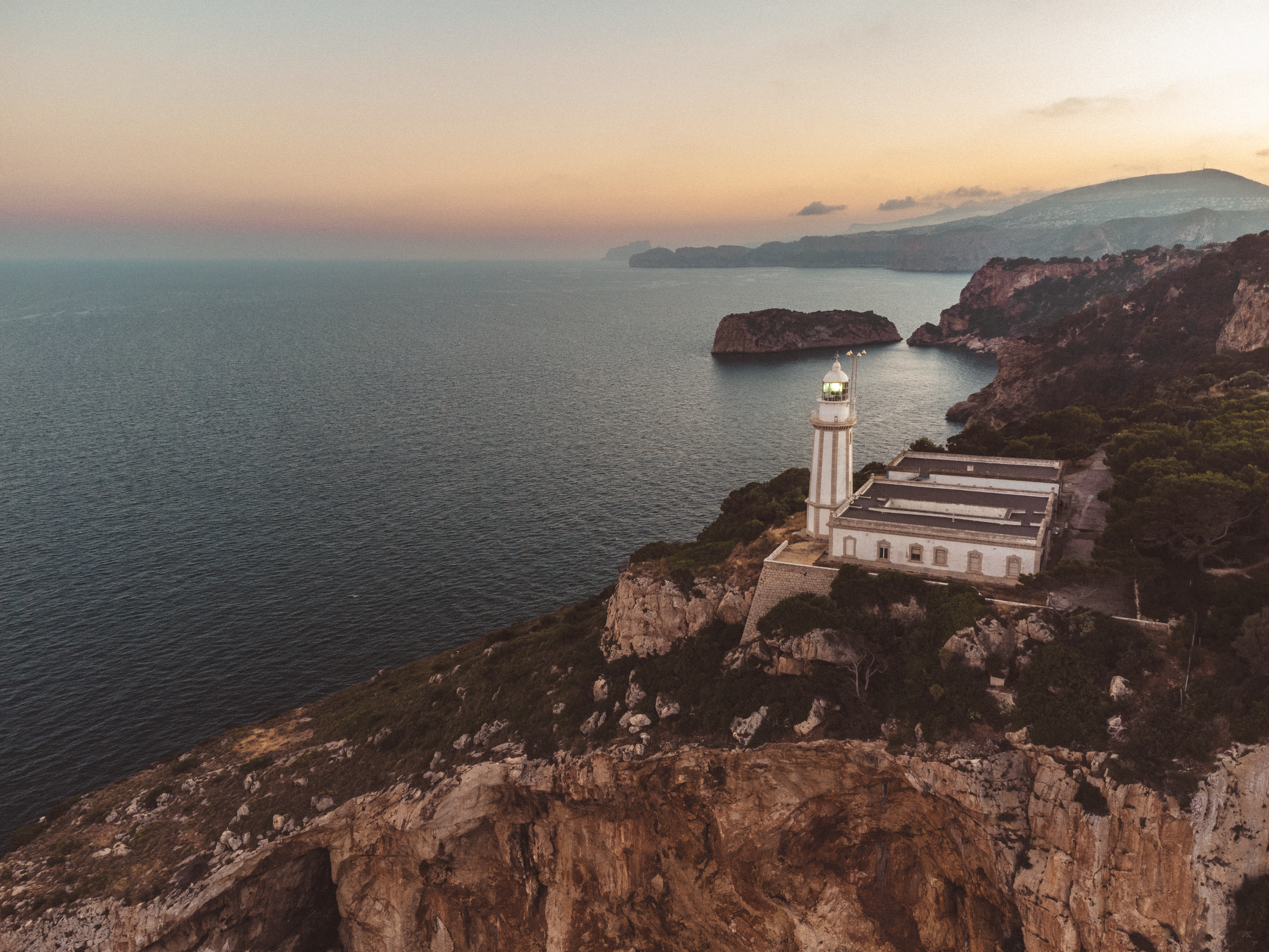 A lighthouse on a rocky cliff overlooking the ocean at sunset with distant mountains in the background.