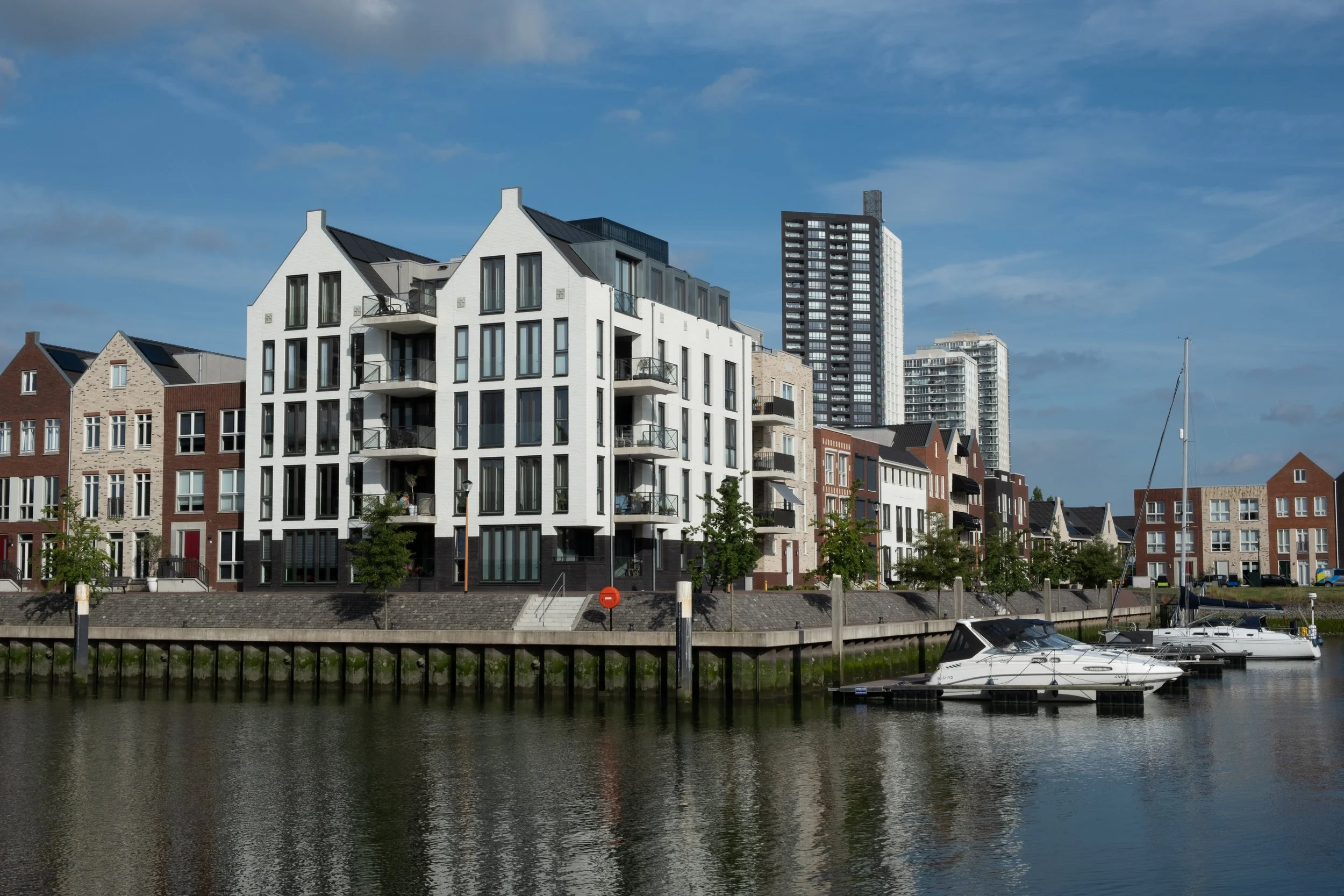 Modern white and brick residential buildings along a waterfront with boats docked nearby and a blue sky overhead.