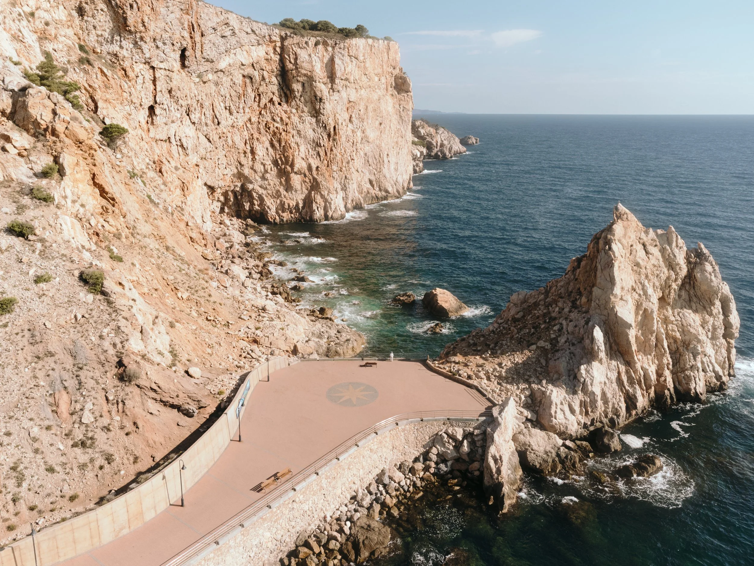 A coastal cliffs with a walkway at the base, overlooking the ocean. Large rocks and cliffs extend into the water, with waves crashing against them.