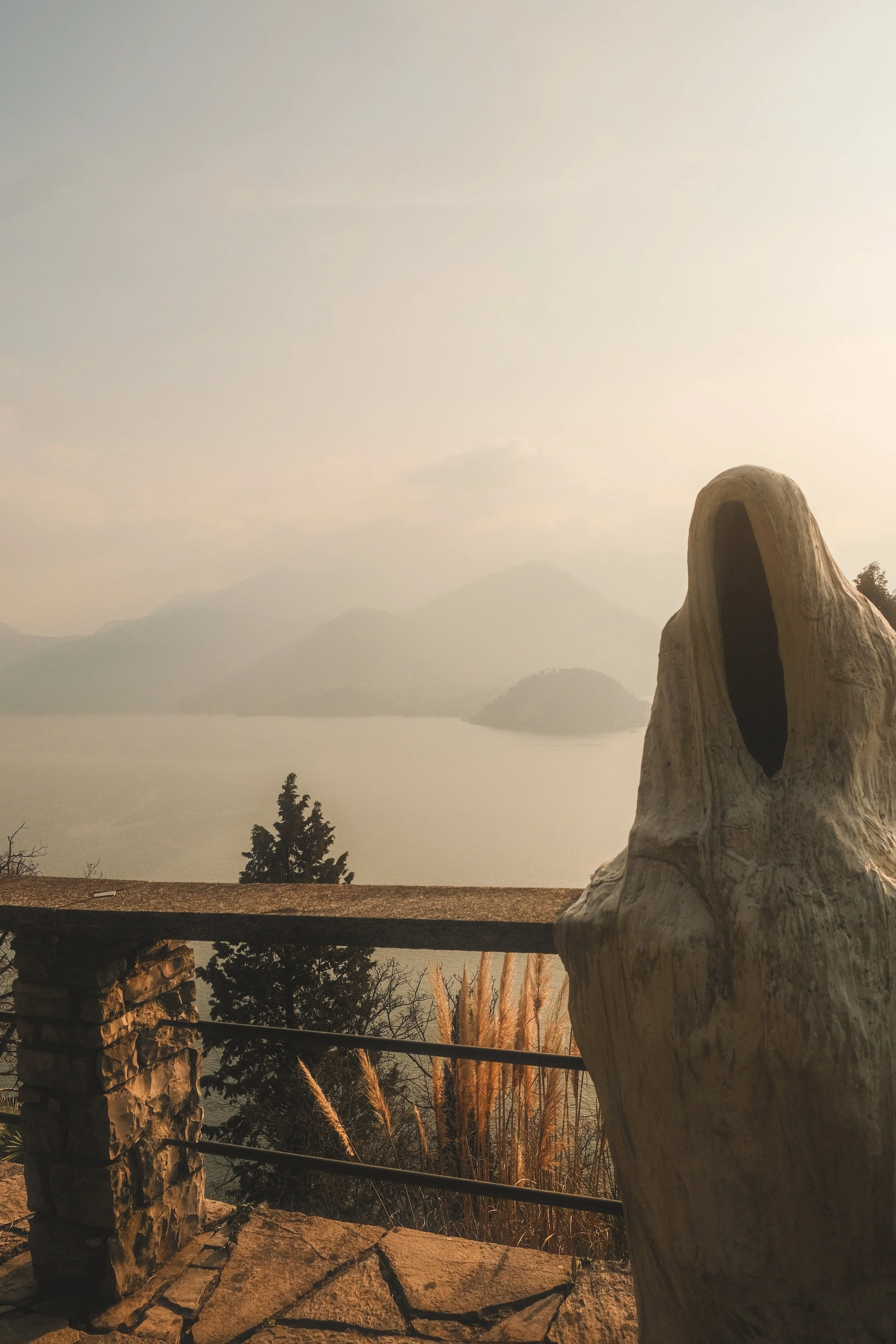 View of a lake or river with distant mountains, a wooden railing, a stone pillar, and a worn wooden sculpture with a hollowed-out face in the foreground.