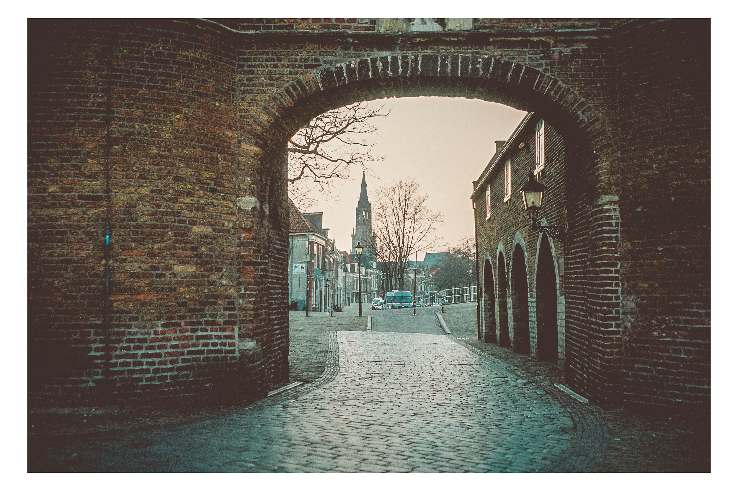 A cobblestone street viewed through a brick archway, with a church steeple and leafless trees in the background, and historic buildings lining the street.