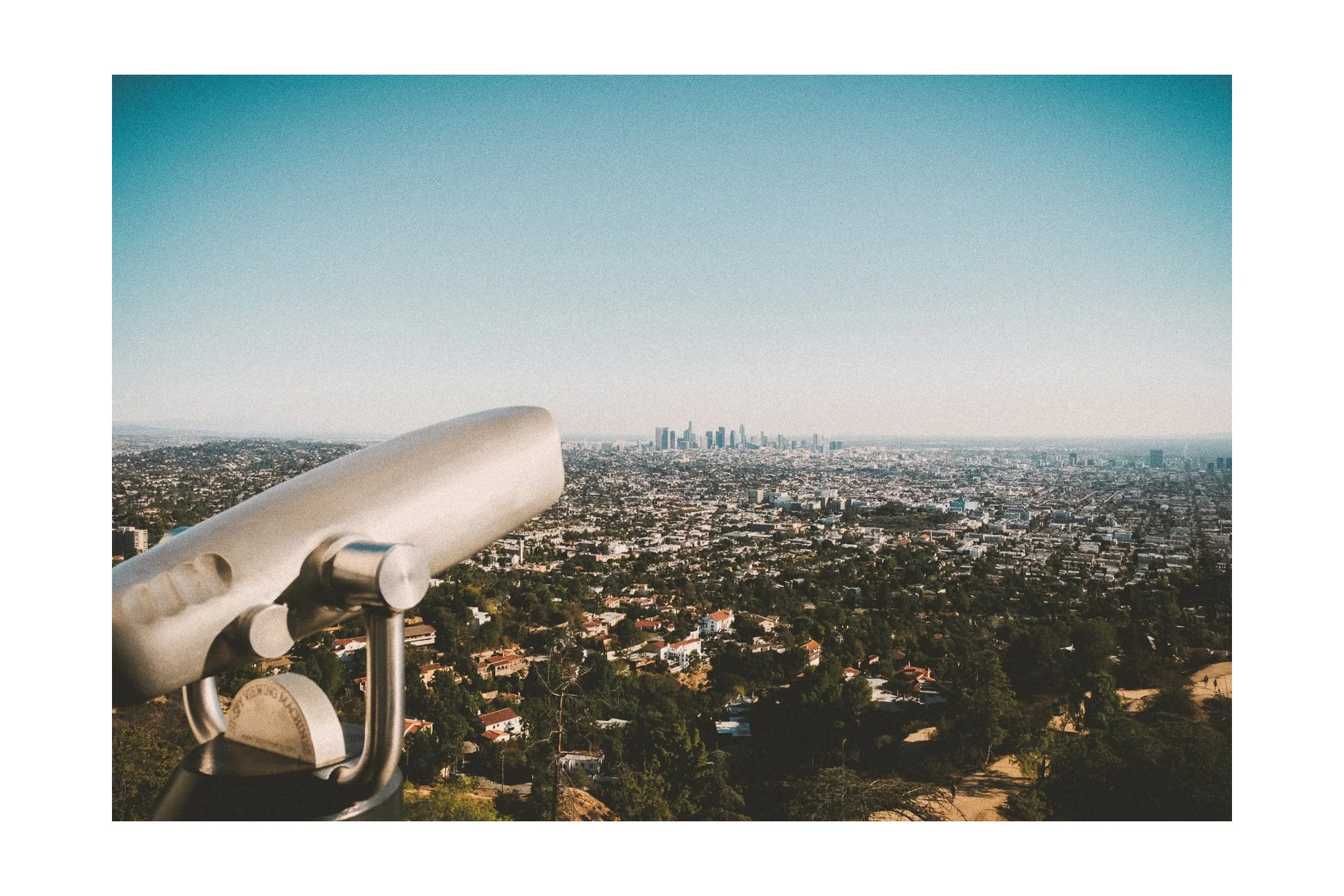 City skyline viewed from a hilltop observation point with a telescope in the foreground, overlooking densely packed city buildings and greenery.