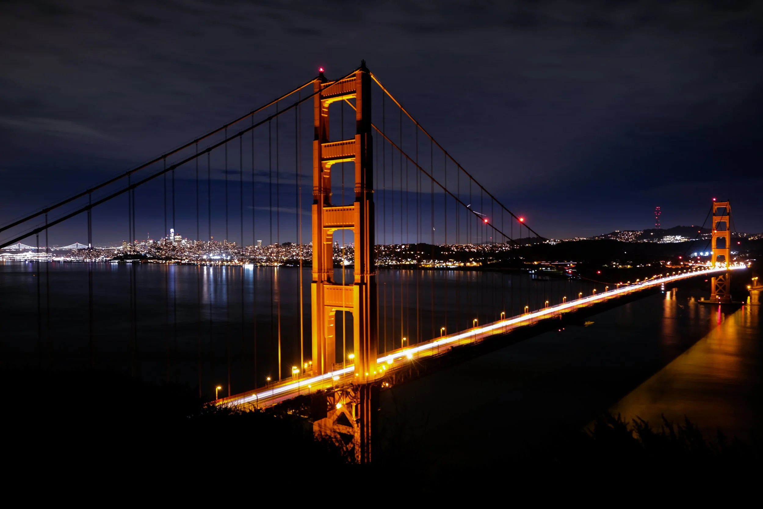 Night view of the Golden Gate Bridge in San Francisco illuminated with orange lights, with city lights and skyline in the background.
