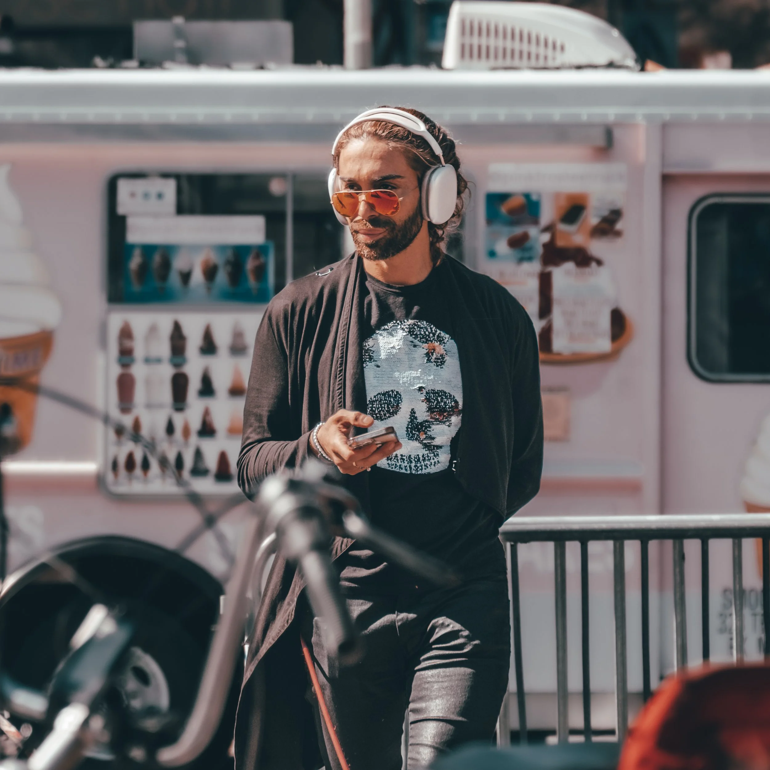 A man wearing sunglasses and large over-ear headphones stands near an ice cream truck, looking at his phone.