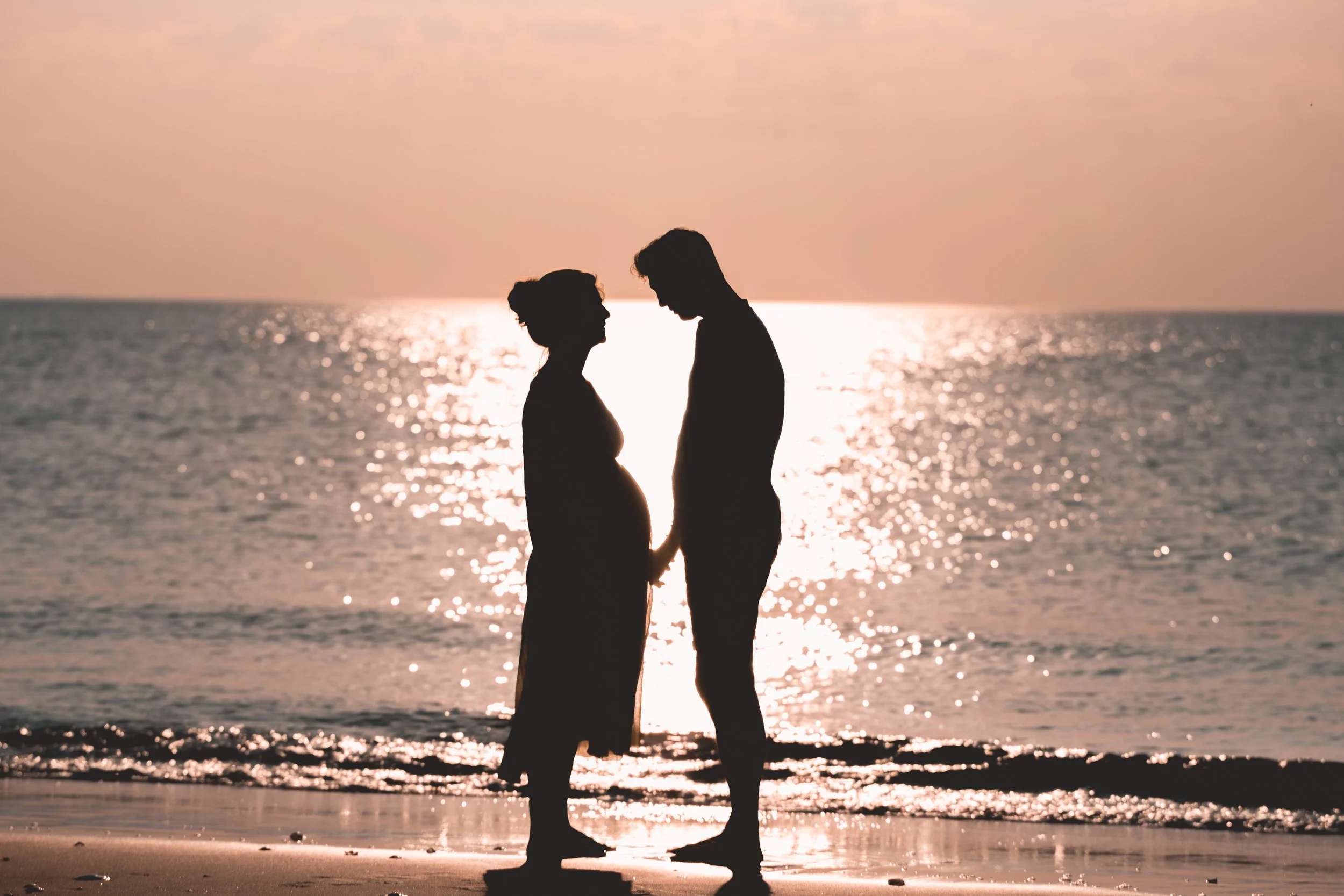 Silhouetted couple holding hands on a beach at sunset, facing each other with the ocean and shimmering water in the background.