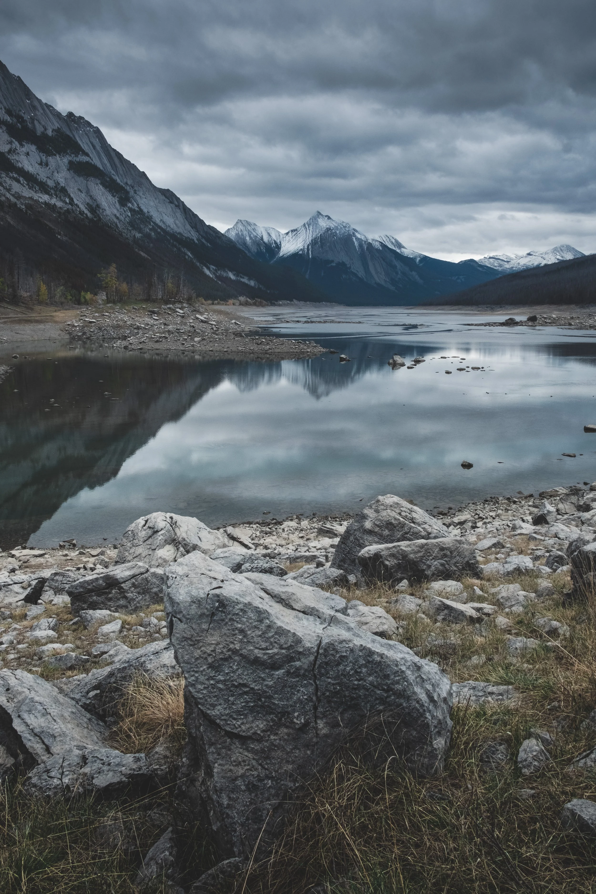 Mountain landscape with snow-capped peaks, a calm reflective lake, and rocky foreground under a cloudy sky.