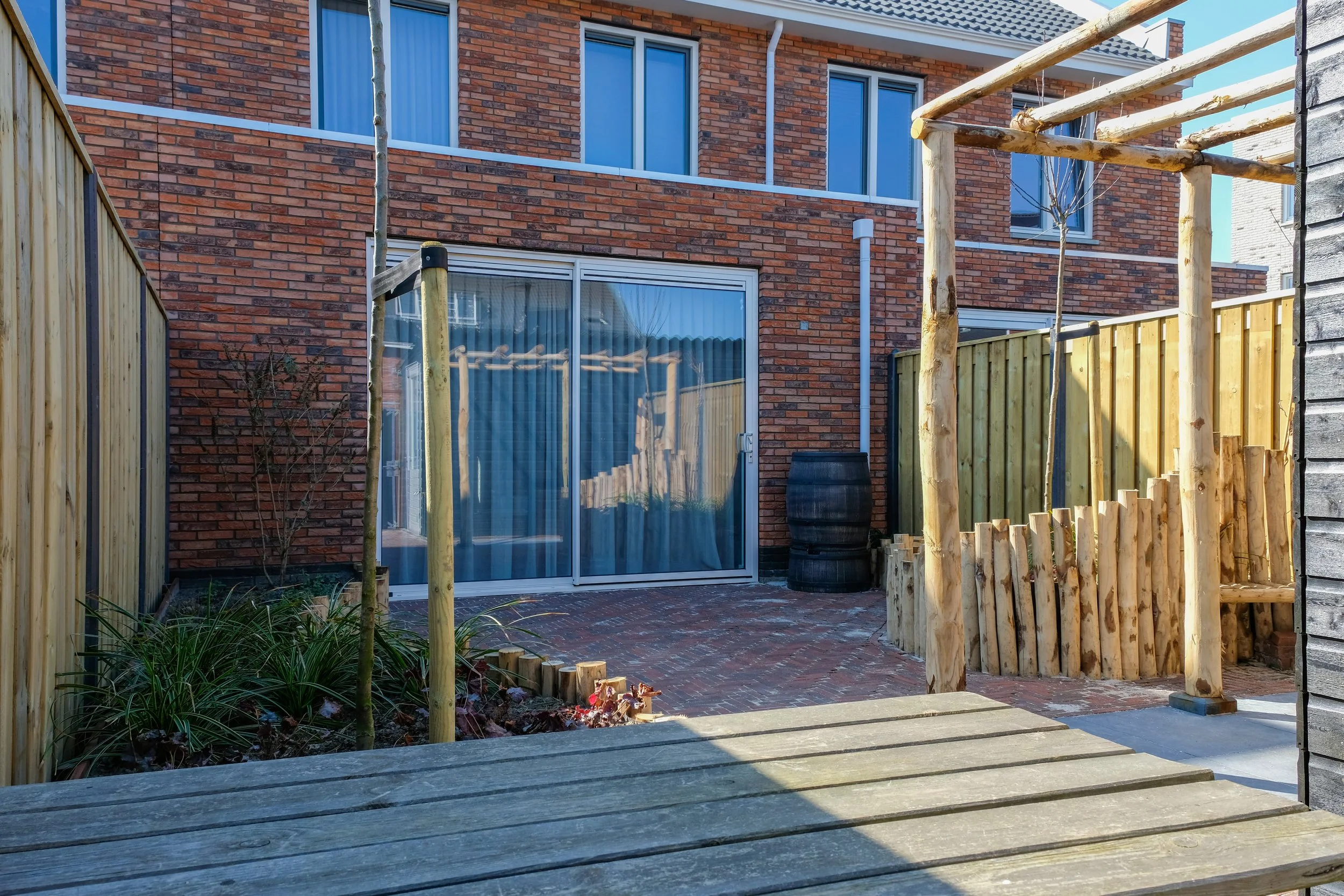 Backyard patio area with brick wall, sliding glass door, young tree, wooden fence, and wooden construction framework.