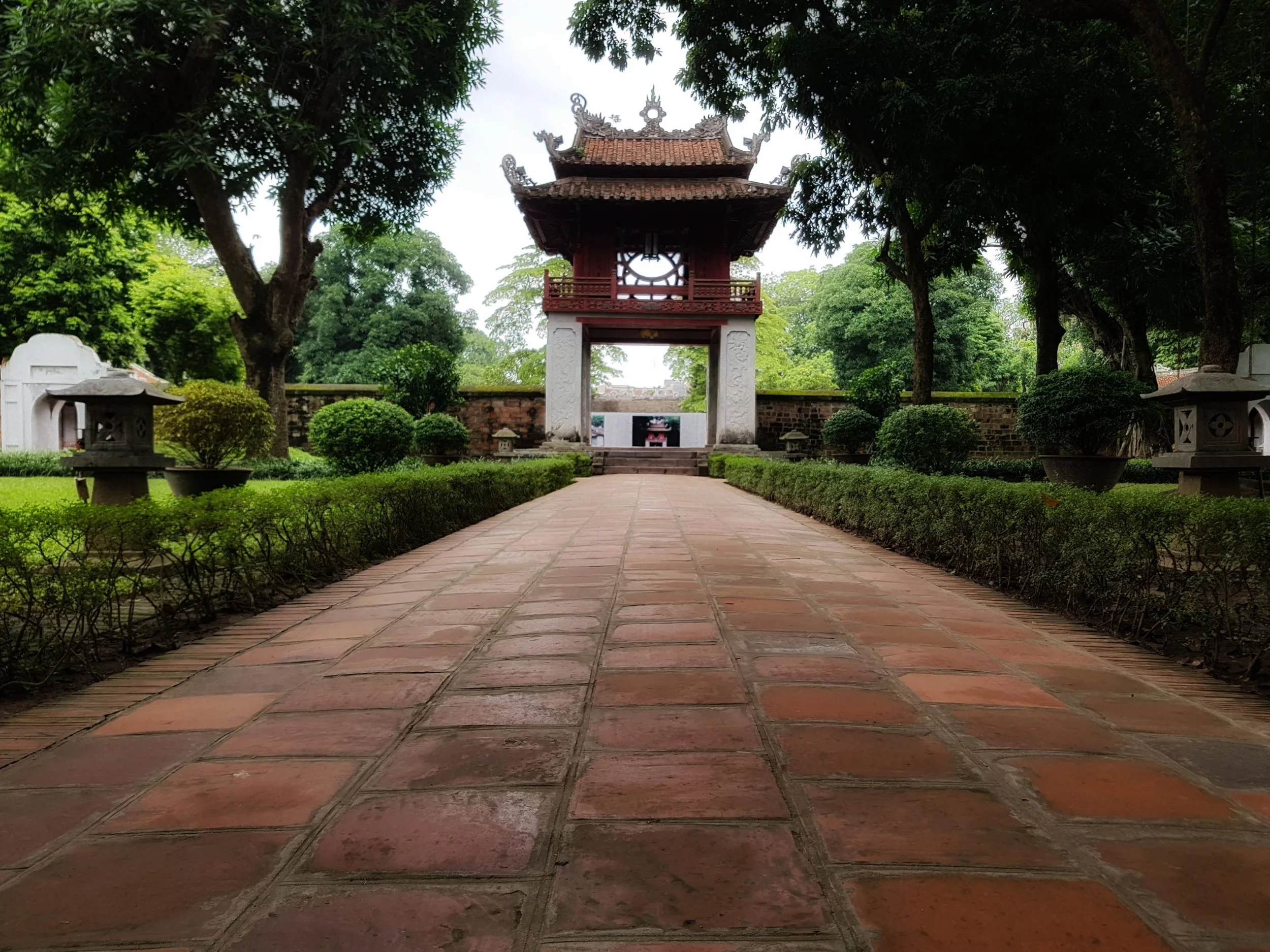 A traditional Asian entrance gate with a tiled roof, framed by lush green trees and neatly trimmed bushes, situated along a brick pathway in a serene garden setting.
